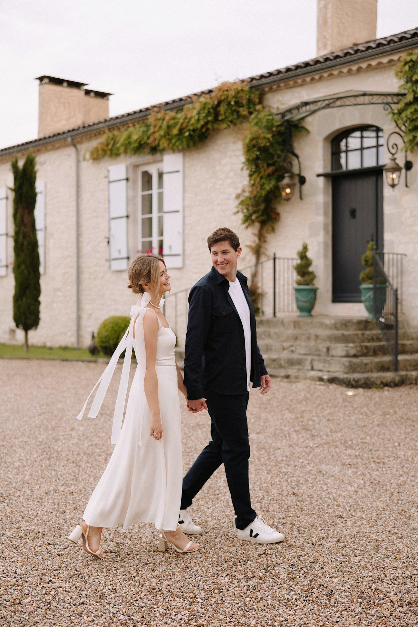 A woman in a white dress and a man in dark pants and a jacket walk hand in hand outside a light stone building with white shutters and greenery.