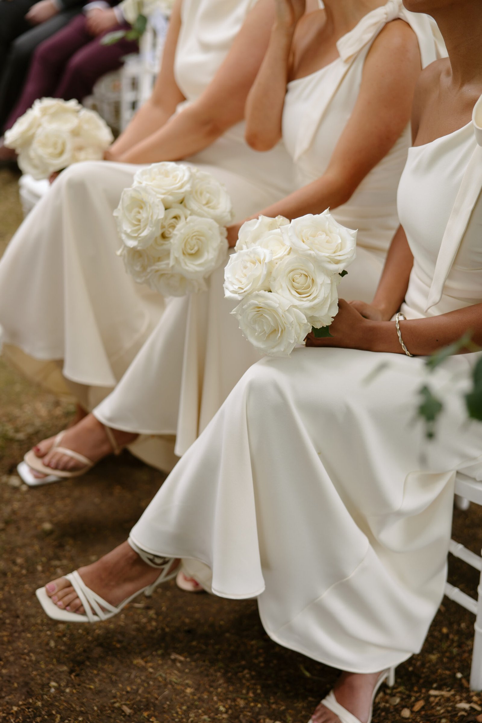 Three women in white dresses sit side by side, each holding a bouquet of white roses. Only their lower bodies and bouquets are visible.