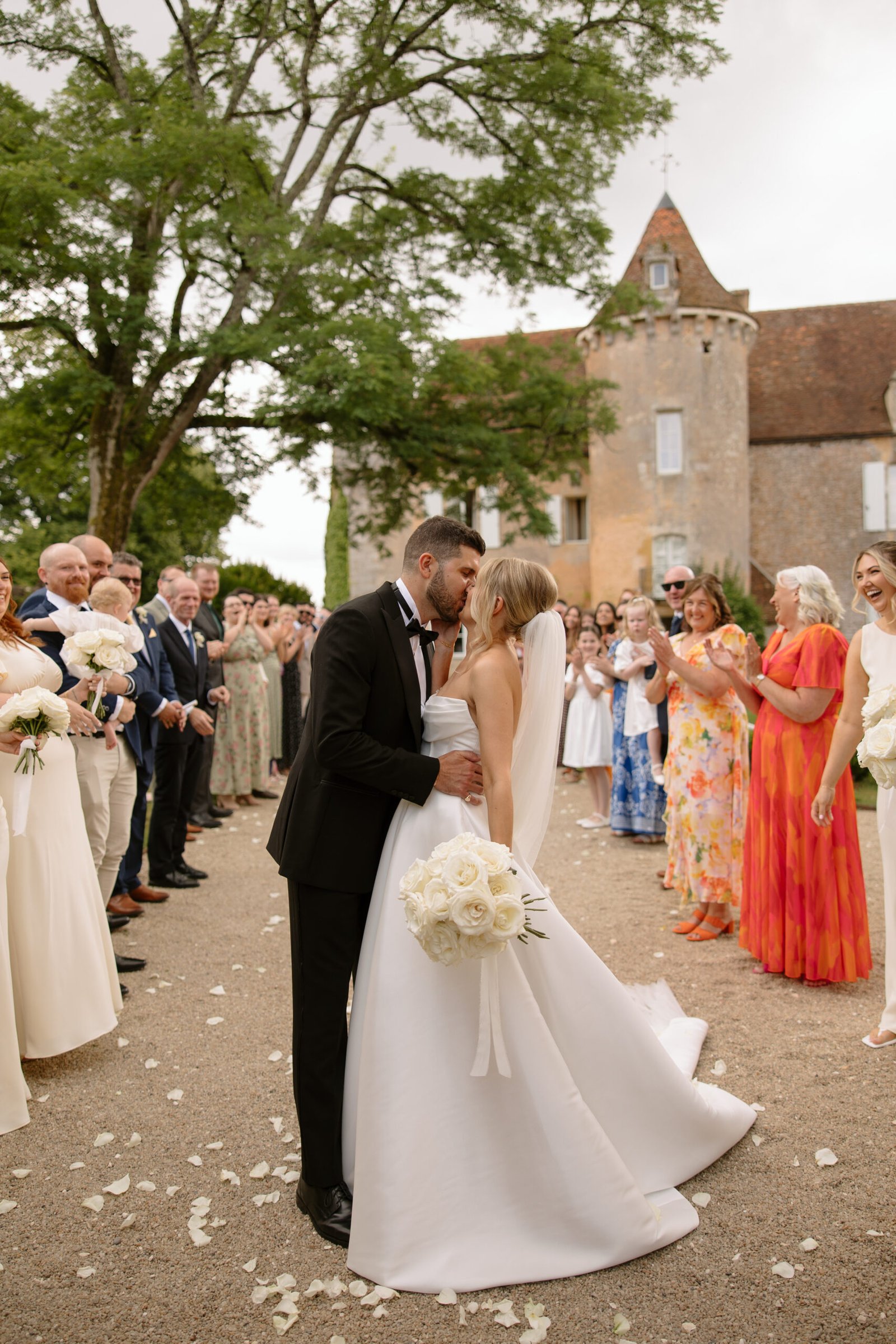 A bride and groom kiss outdoors among guests, with a stone building and trees in the background; the bride holds a bouquet of white roses.