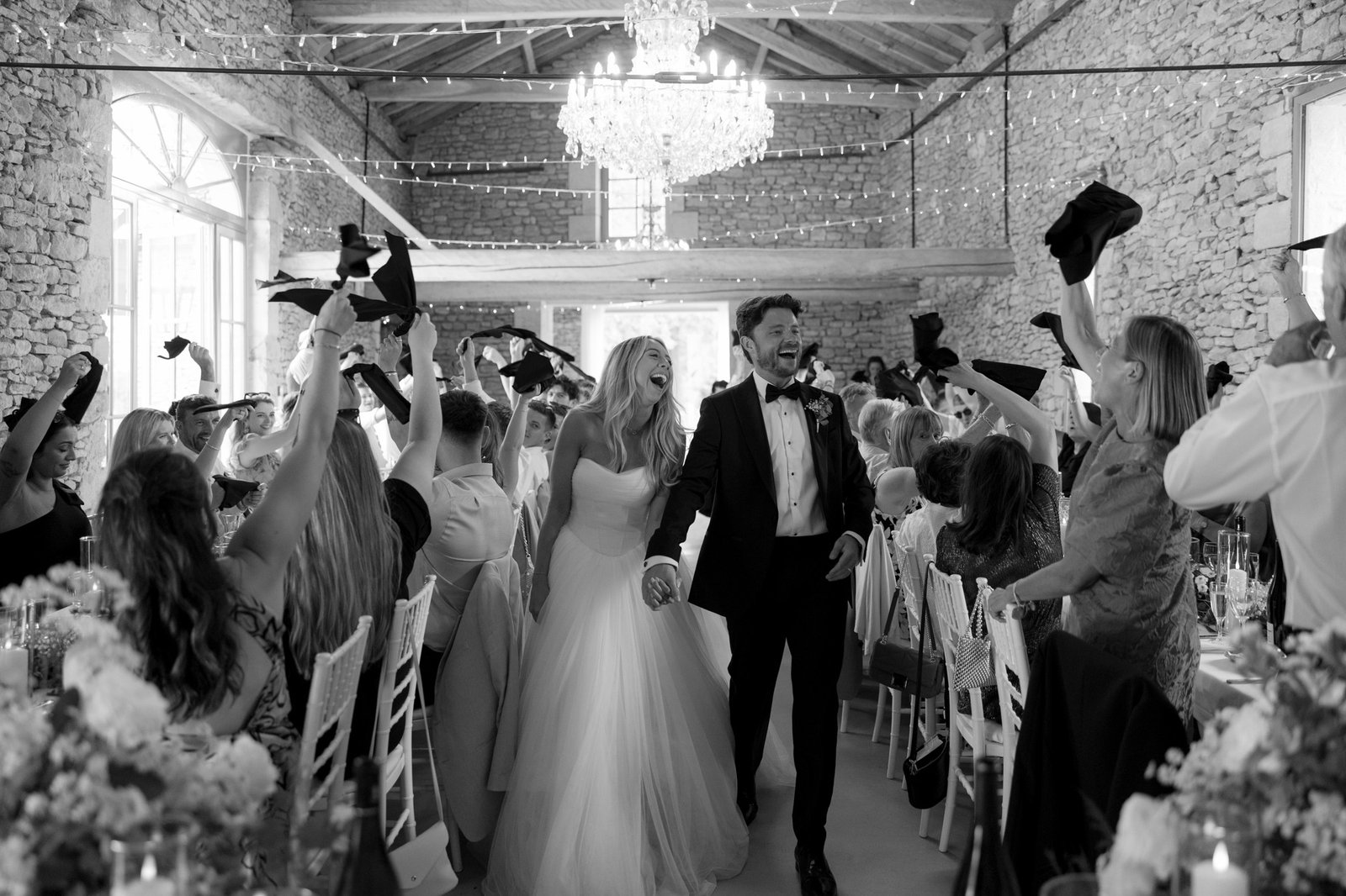 A bride and groom walk hand in hand down the aisle in a stone-walled hall as guests cheer and wave napkins in celebration.