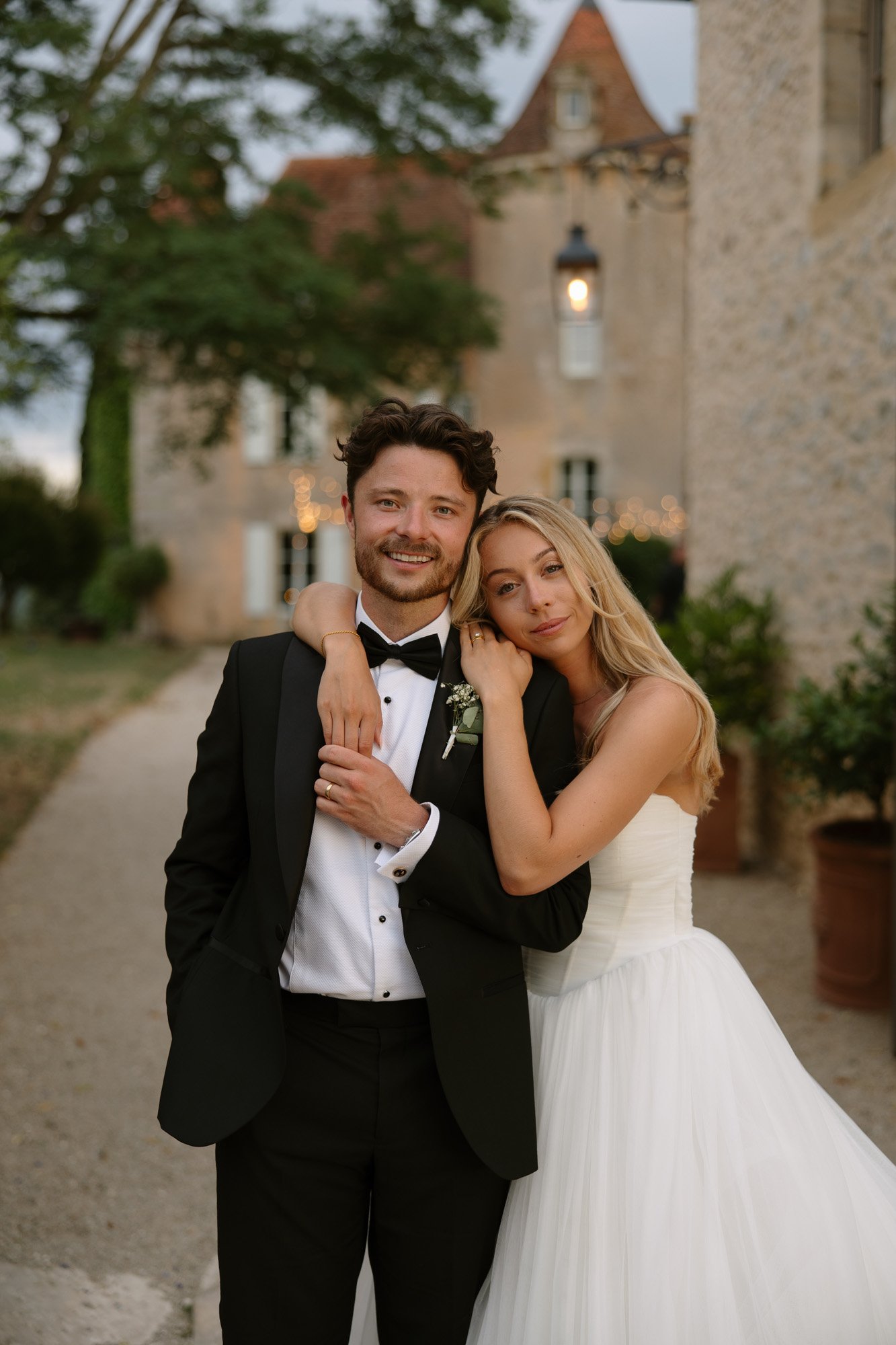 A bride in a white gown stands with her arm around a groom in a black tuxedo, both smiling, outside a stone building with greenery.