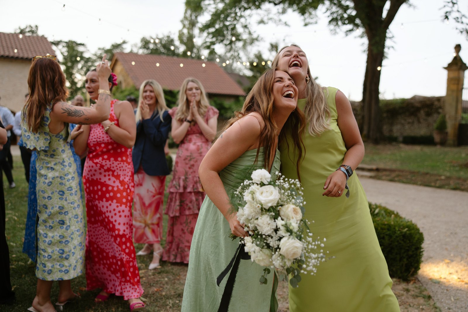 Two women in pastel dresses laugh and hold a bouquet of flowers outdoors, while a group of women in colorful dresses applaud and celebrate in the background.