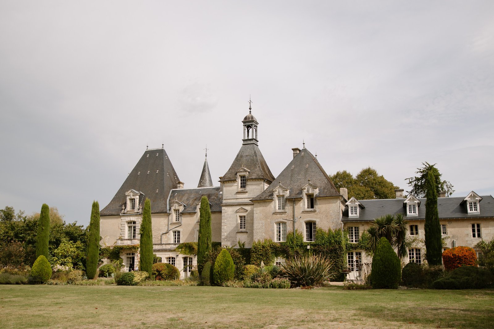 A stone chateau with multiple peaked roofs and towers, surrounded by trees, tall shrubs, and a grassy lawn under a cloudy sky.