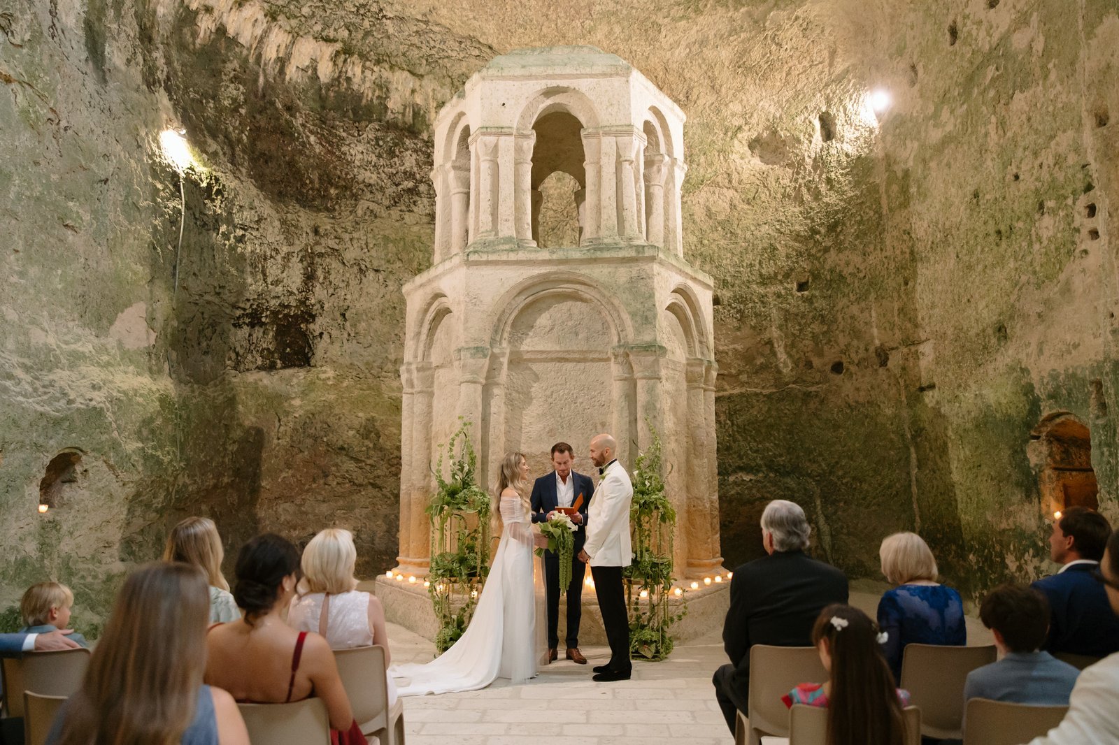 A couple stands with an officiant during a wedding ceremony in front of an ancient stone structure, while guests are seated and watching. Aubeterre sur dronne wedding.