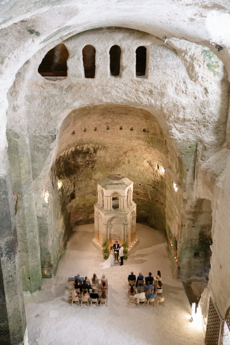 A small group of people attend a wedding ceremony inside an ancient, cave-like stone chamber with high vaulted ceilings and arched windows.