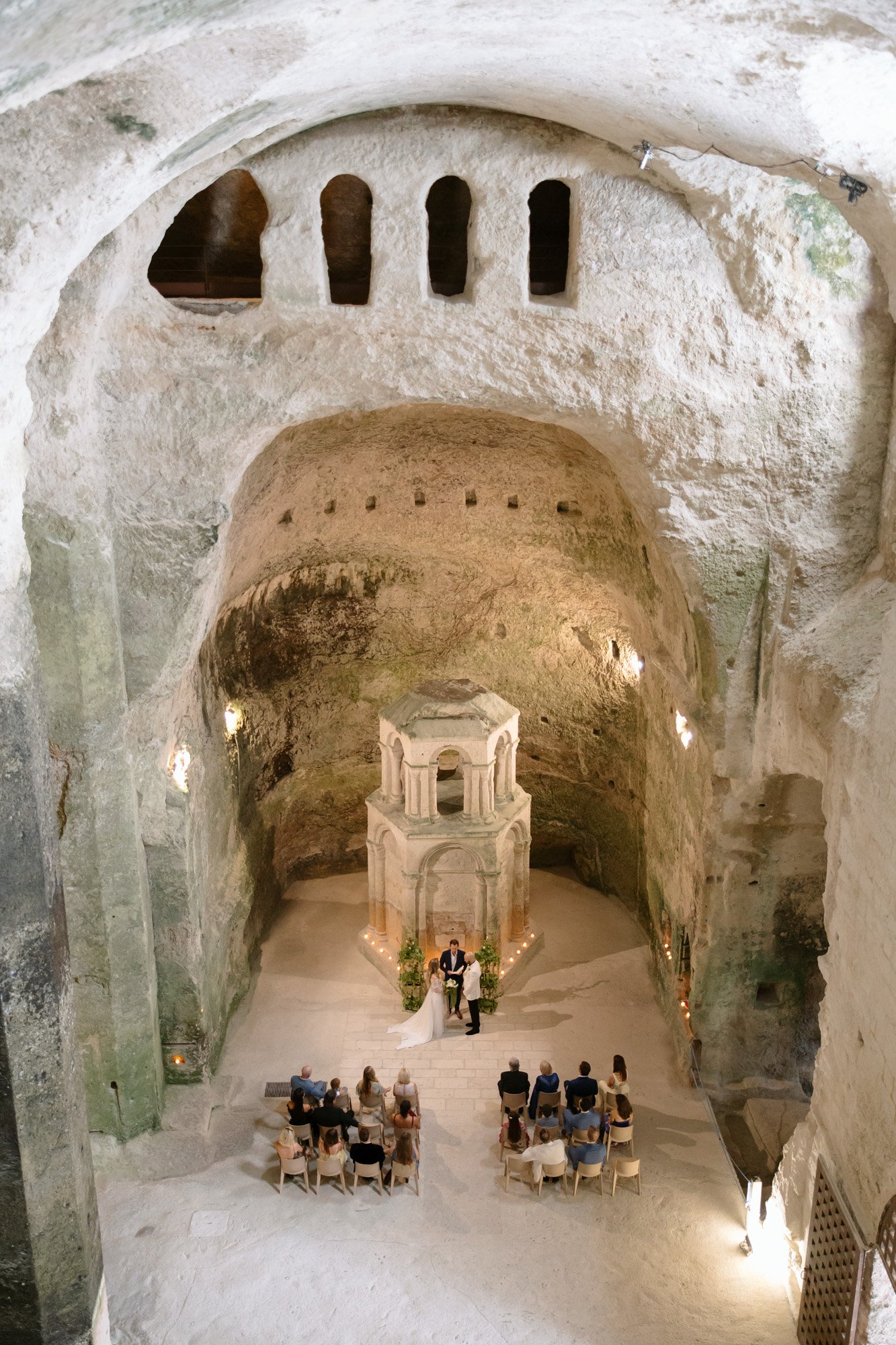 A small group of people attend a wedding ceremony inside an ancient, cave-like stone chamber with high vaulted ceilings and arched windows.