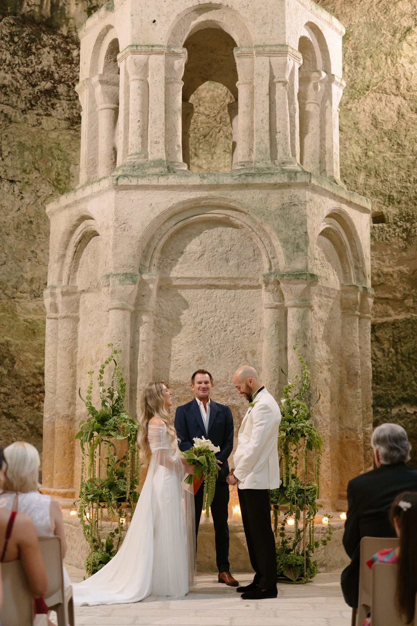 A couple stands before an officiant during a wedding ceremony inside a historic stone structure, surrounded by seated guests.