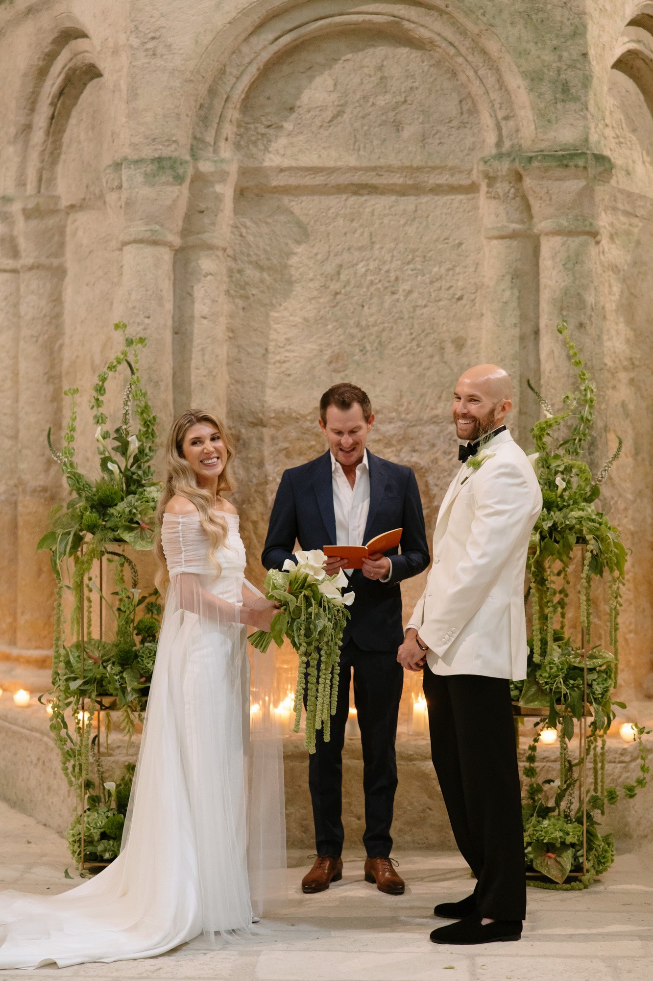 A bride and groom stand facing each other with a smiling officiant between them, surrounded by greenery and candles in a stone-walled venue. Aubeterre sur dronne wedding.