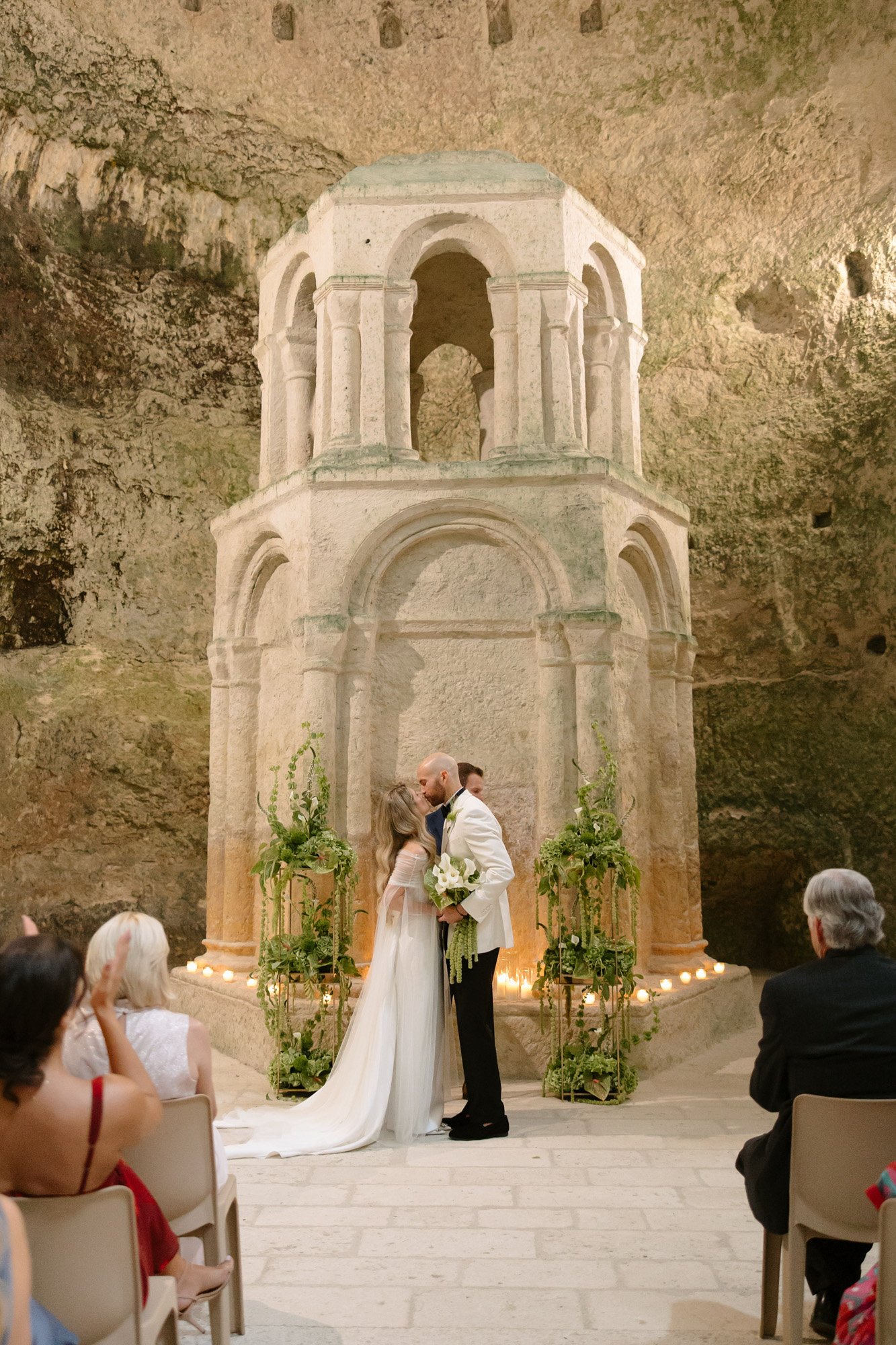 A bride and groom kiss at the altar during their wedding ceremony in front of a stone structure inside a cave-like venue, with guests seated and watching. Aubeterre sur dronne wedding.