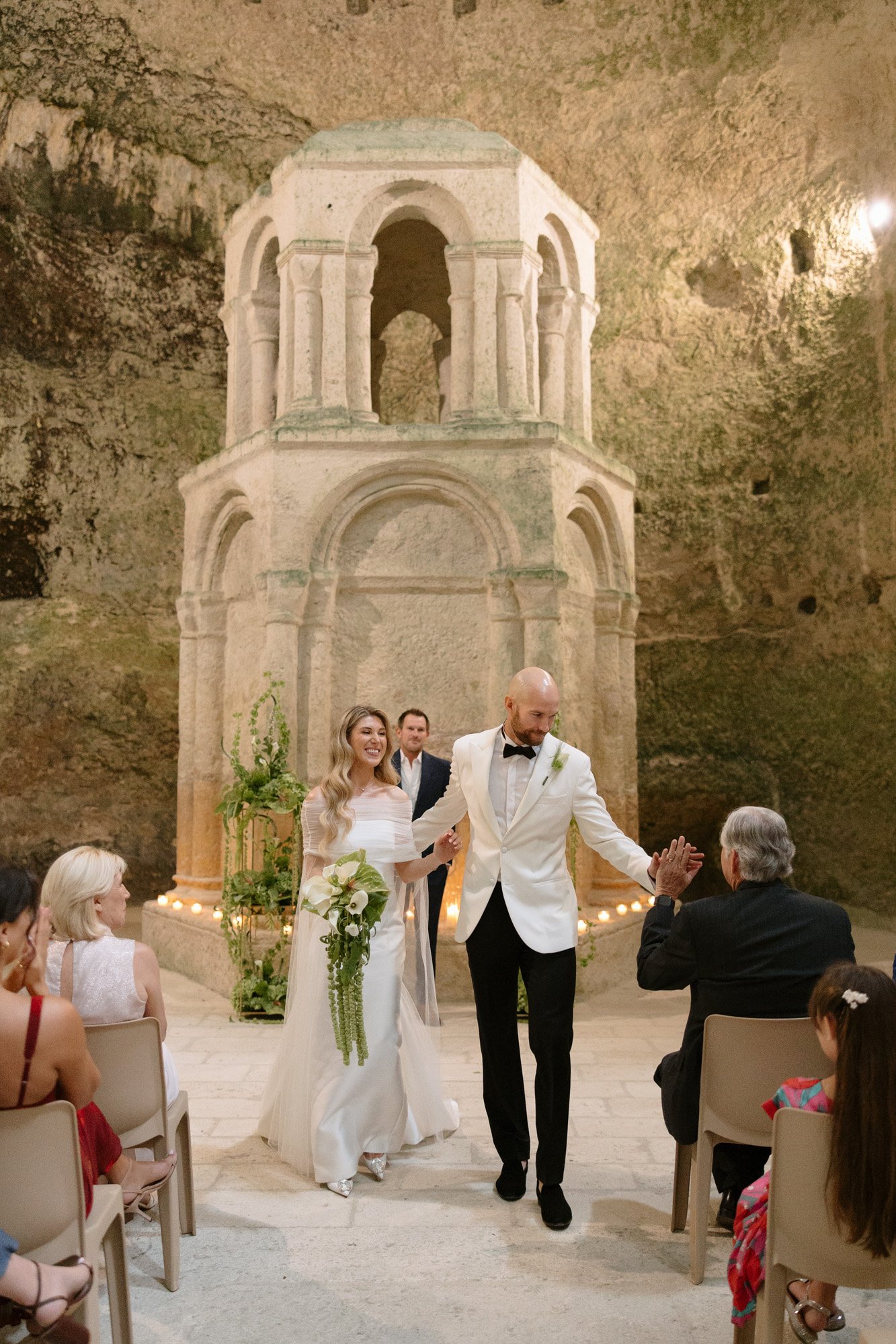 A bride and groom walk down the aisle in an ancient stone chapel, smiling at guests seated on both sides. Aubeterre sur dronne wedding.