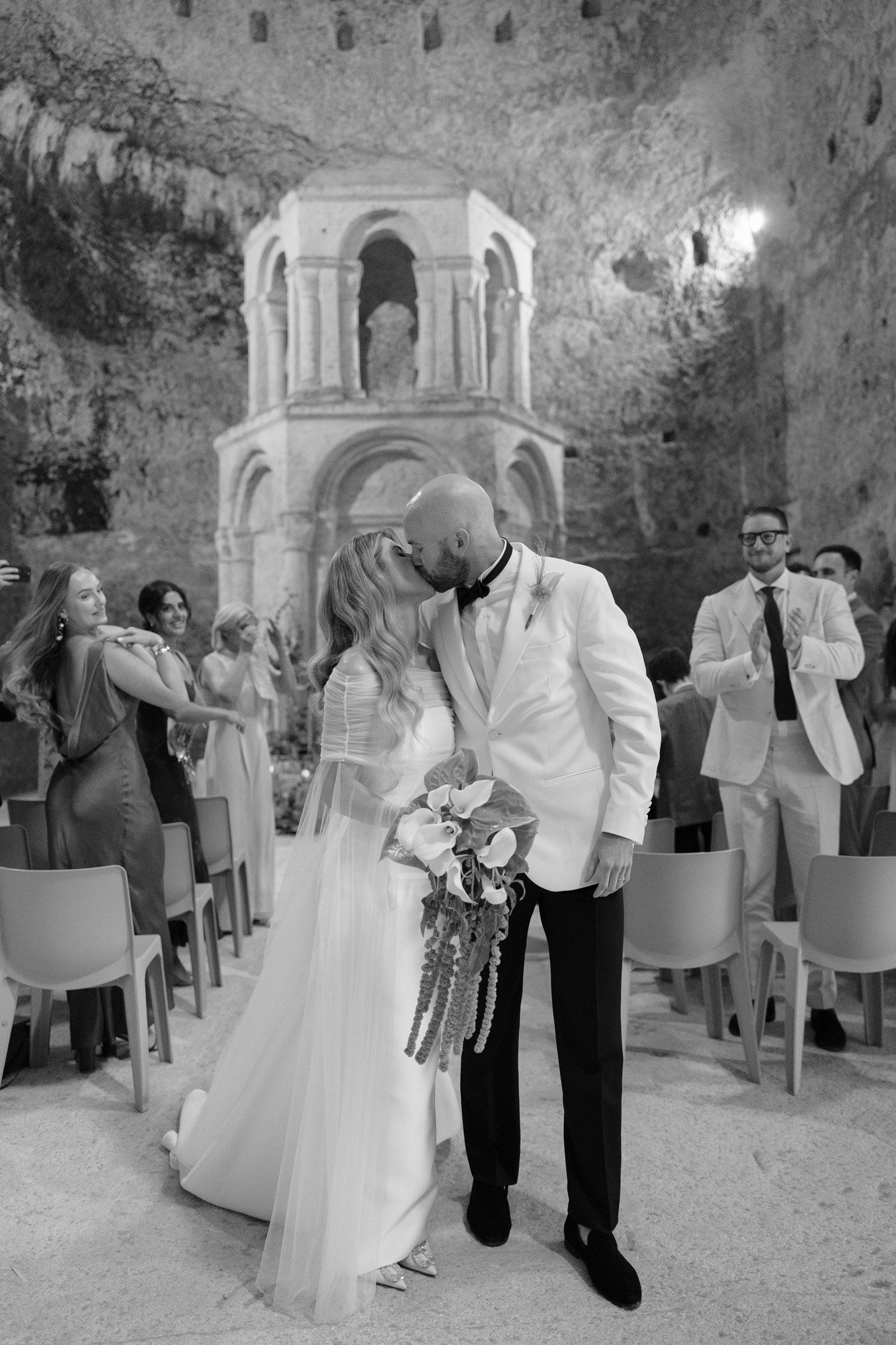 A bride and groom kiss in front of an ancient stone structure while guests applaud in a wedding ceremony setting. Aubeterre sur dronne wedding.