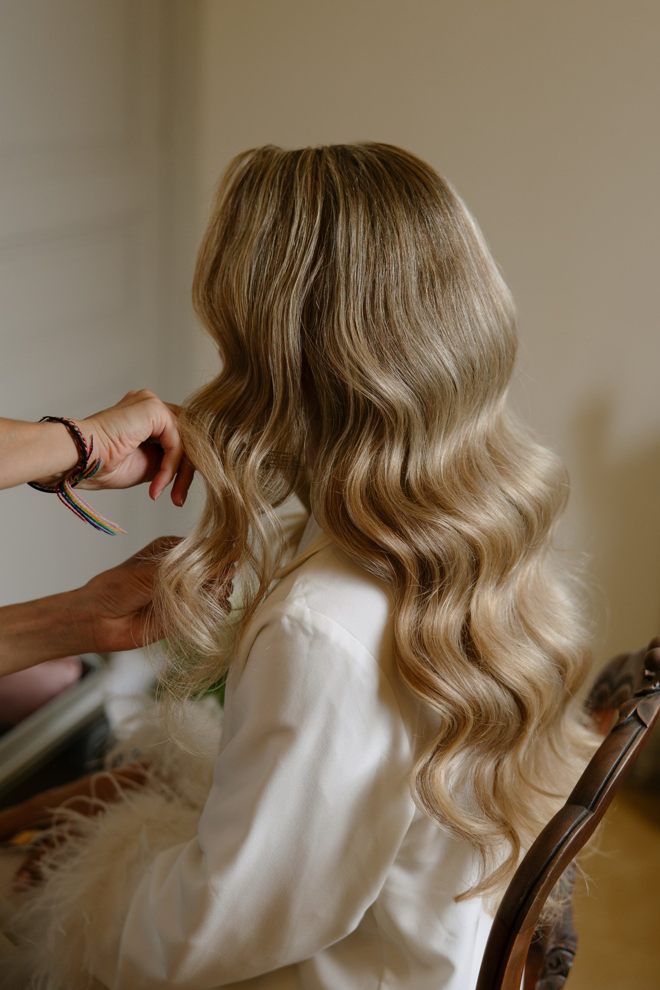 A person with long, wavy blonde hair is seated while another person styles their hair. The setting appears to be indoors.