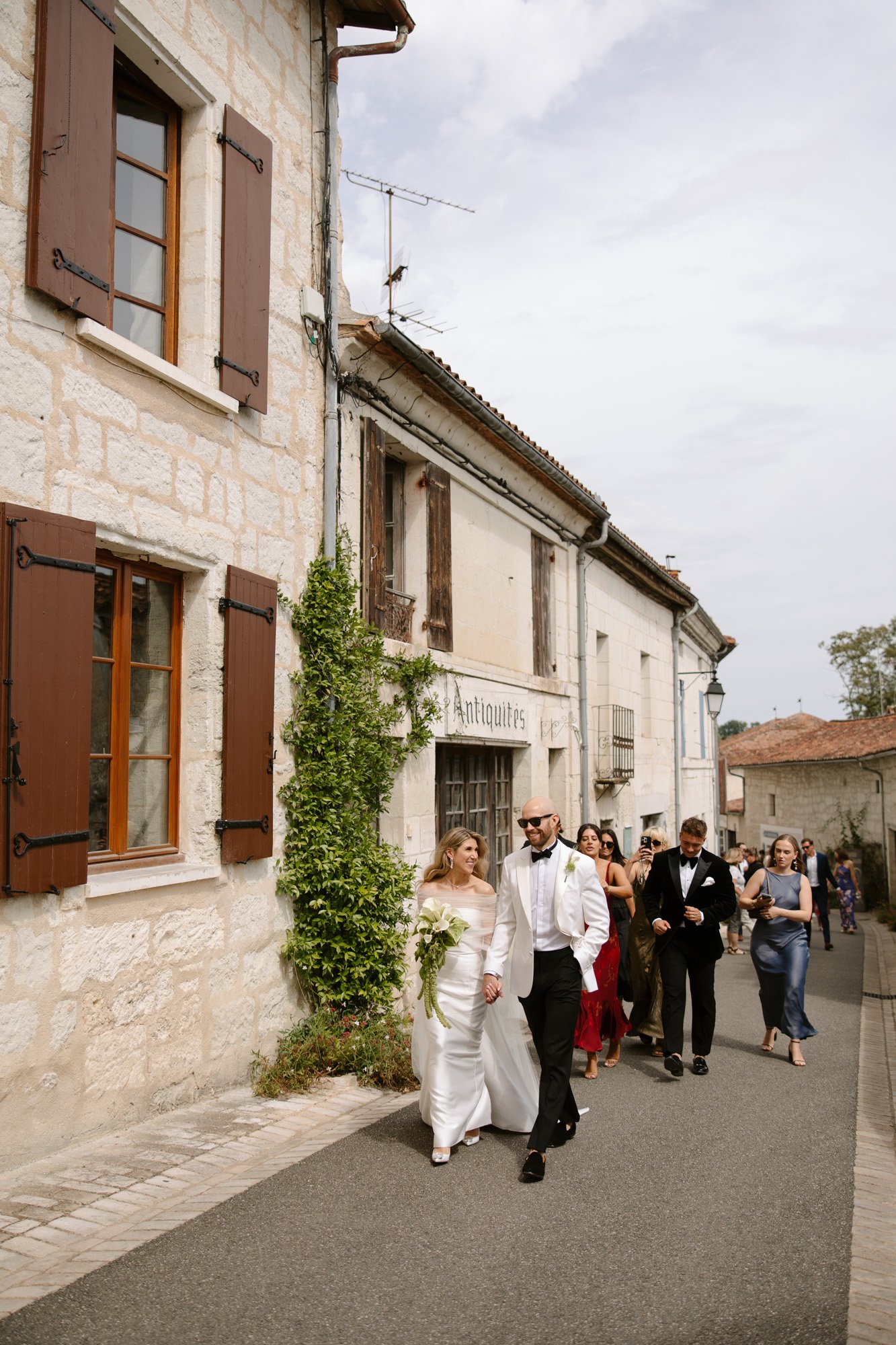 A bride and groom walk down a narrow street with a group of well-dressed guests outside stone buildings on a cloudy day. Aubeterre sur dronne wedding.