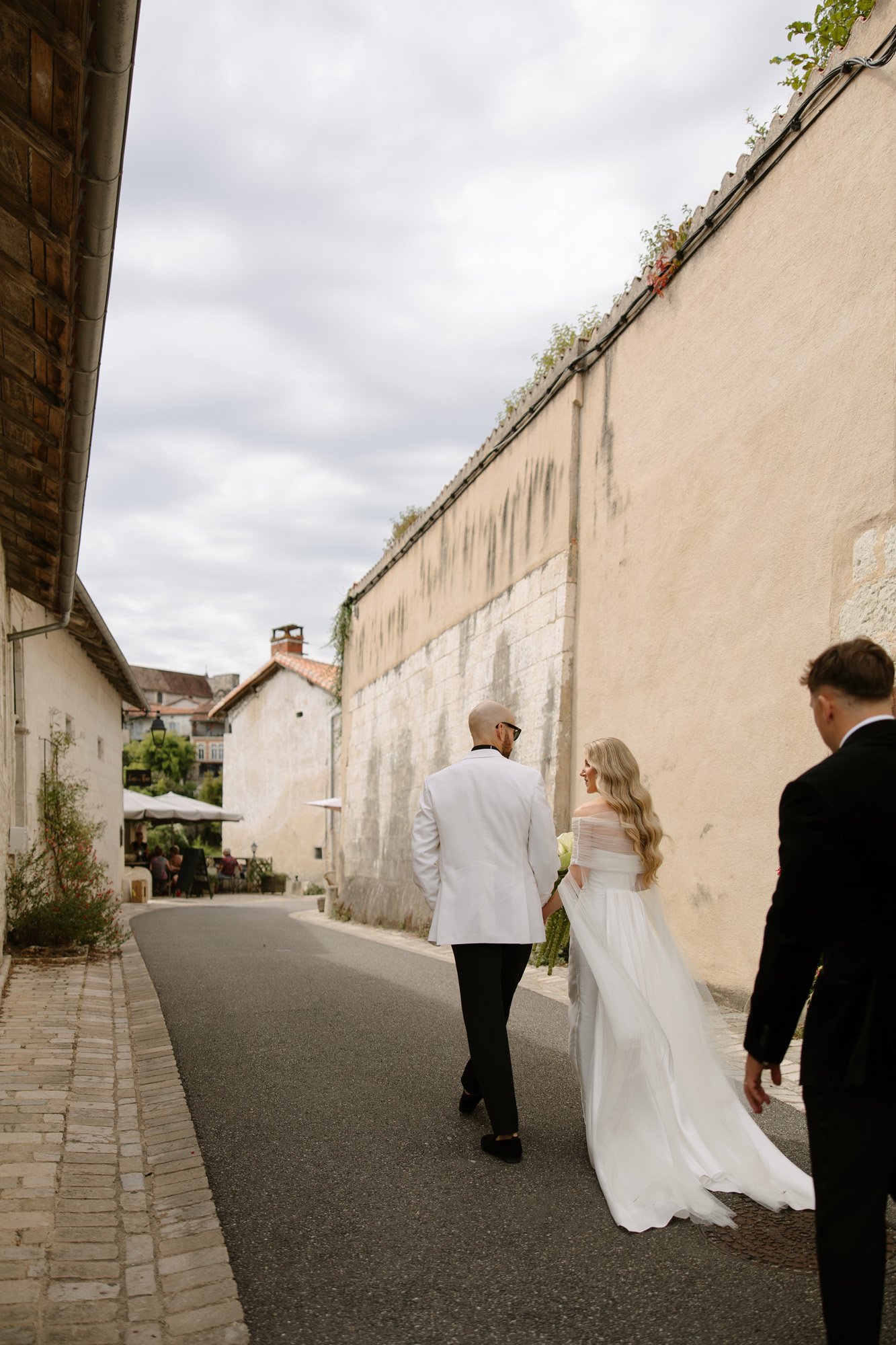 A bride in a white dress and a groom in a white jacket walk down a narrow street, followed by a man in a black suit. The scene is outdoors beside stone buildings. Aubeterre sur dronne wedding.