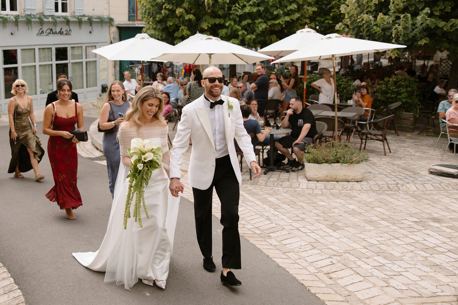 A bride and groom walk hand in hand on a cobblestone street, followed by guests, with outdoor café seating and people in the background. Aubeterre sur dronne wedding.