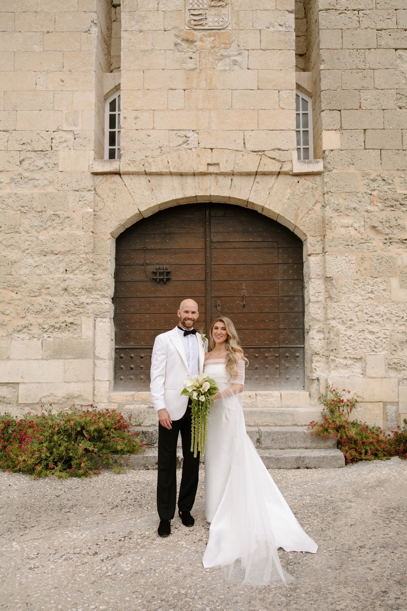 A couple dressed in formal wedding attire stands in front of a large historic stone building with a wooden arched door. Aubeterre sur dronne wedding.