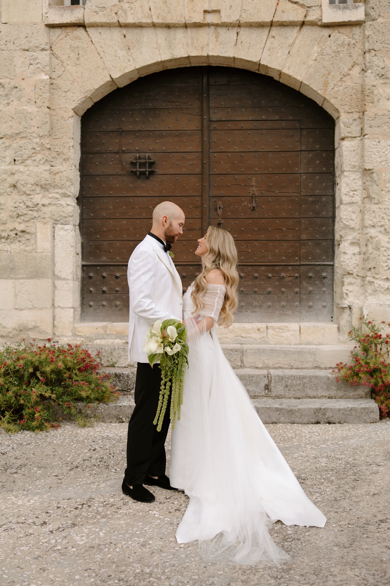A bride and groom stand facing each other outside in front of a large wooden door, dressed in formal wedding attire and holding a bouquet.