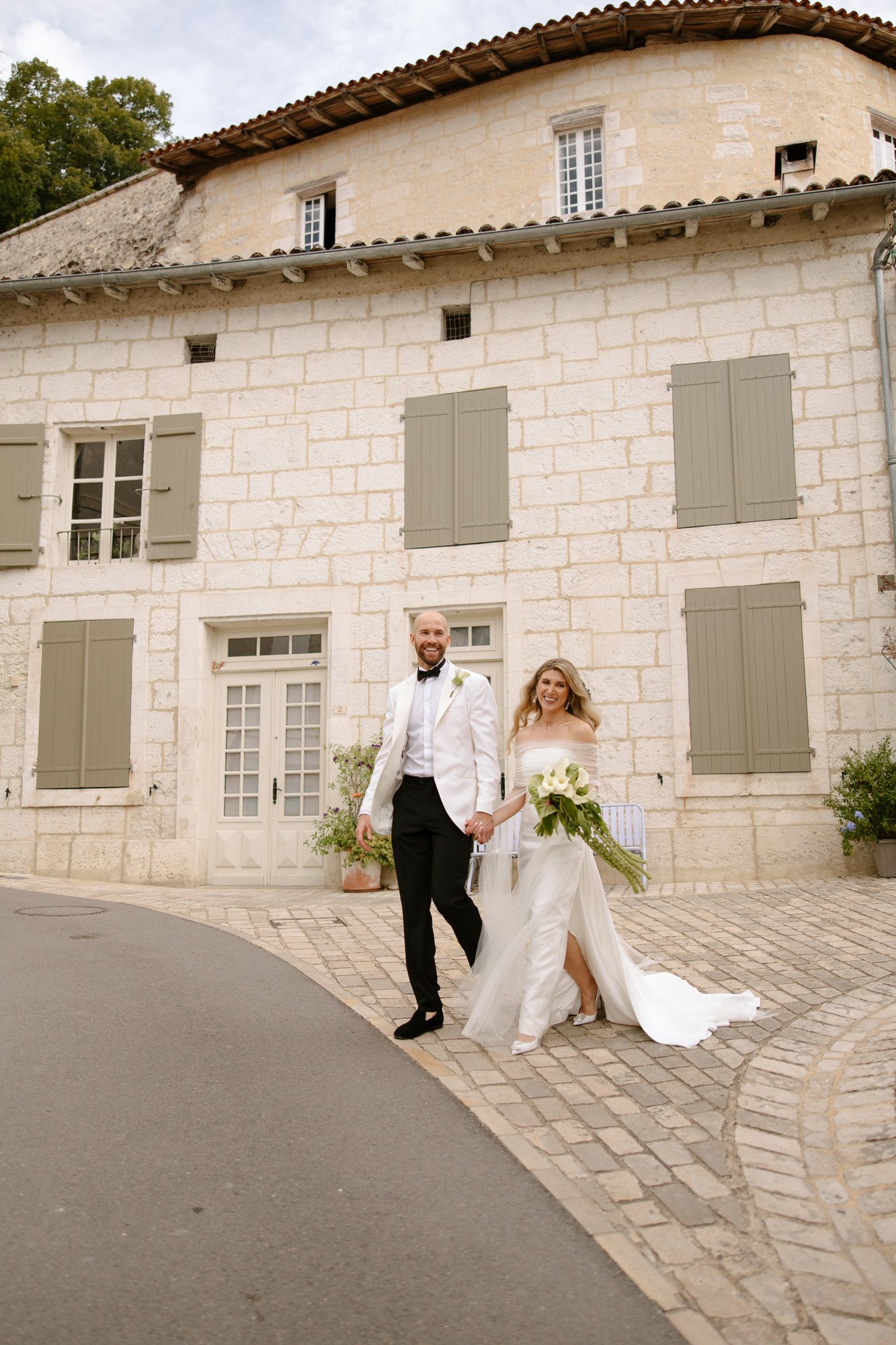 A bride and groom, dressed in formal wedding attire, walk hand in hand on a cobblestone path in front of a stone building with shuttered windows. Aubeterre sur dronne wedding.