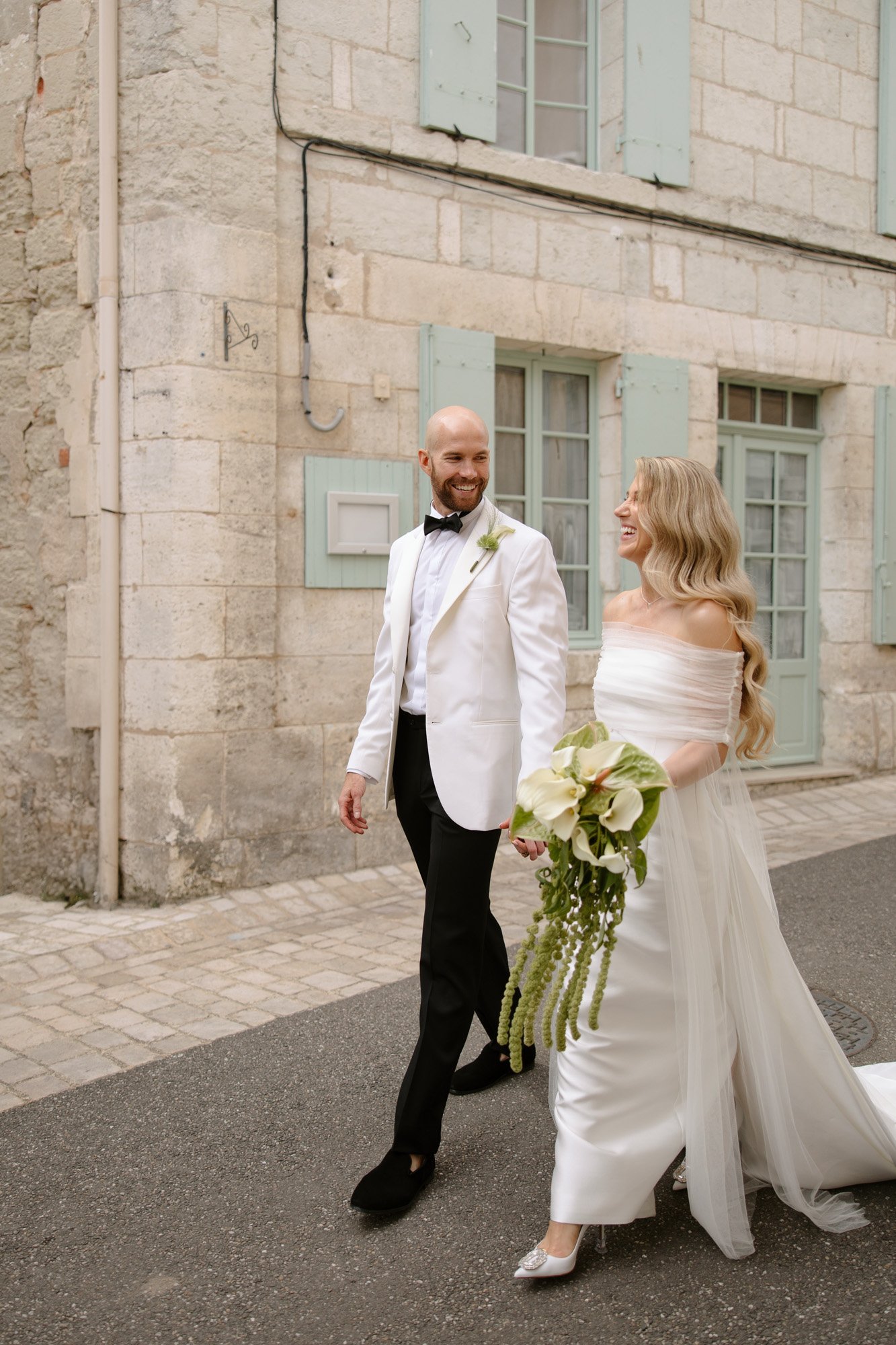 A bride and groom walk hand in hand on a street; the groom wears a white tuxedo jacket and the bride wears an off-shoulder white dress and holds a bouquet. Aubeterre sur dronne wedding.