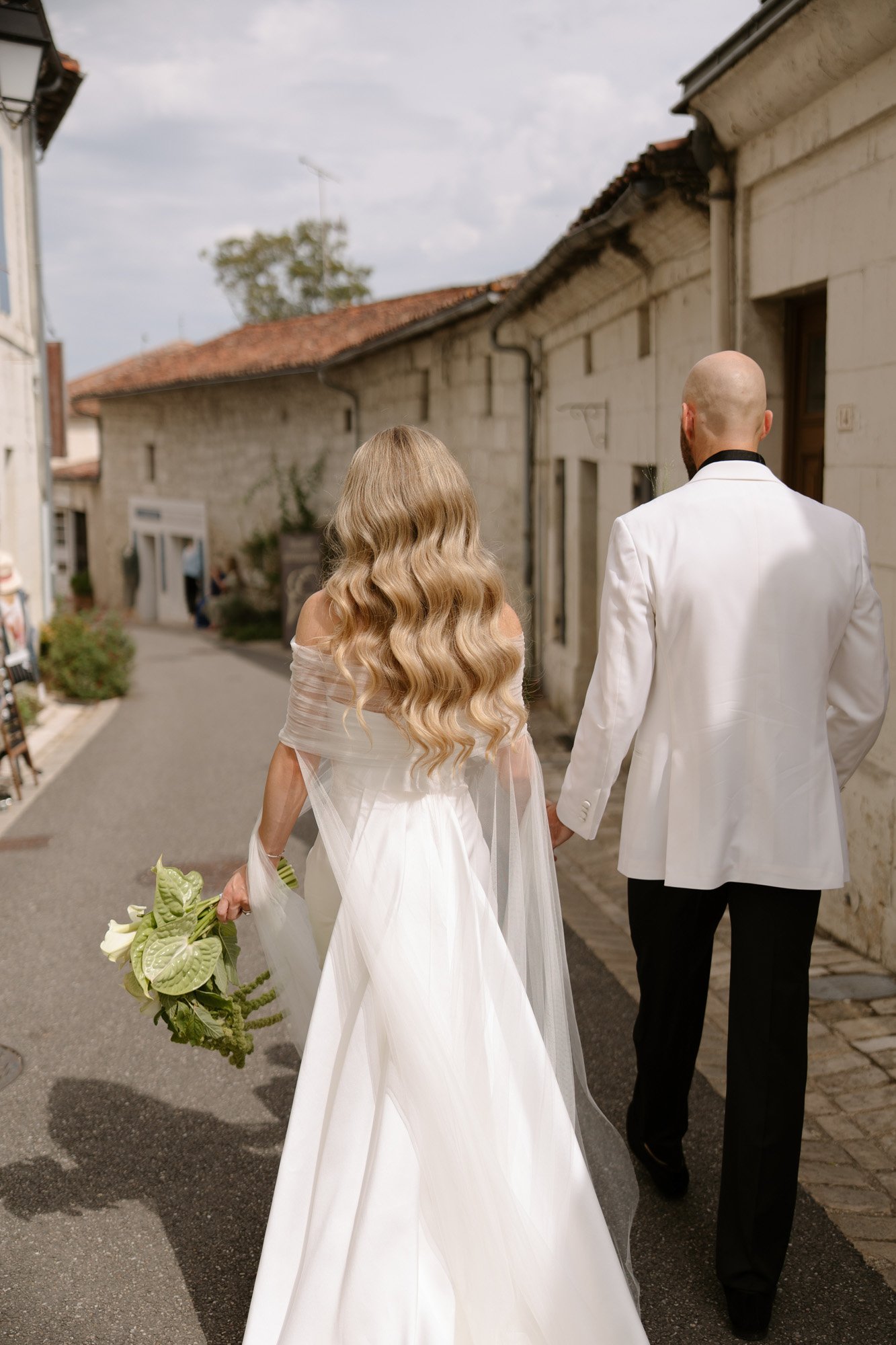 A bride and groom walk down a narrow street, both dressed in white formal attire; the bride holds a bouquet of green and white flowers. Aubeterre sur dronne wedding.