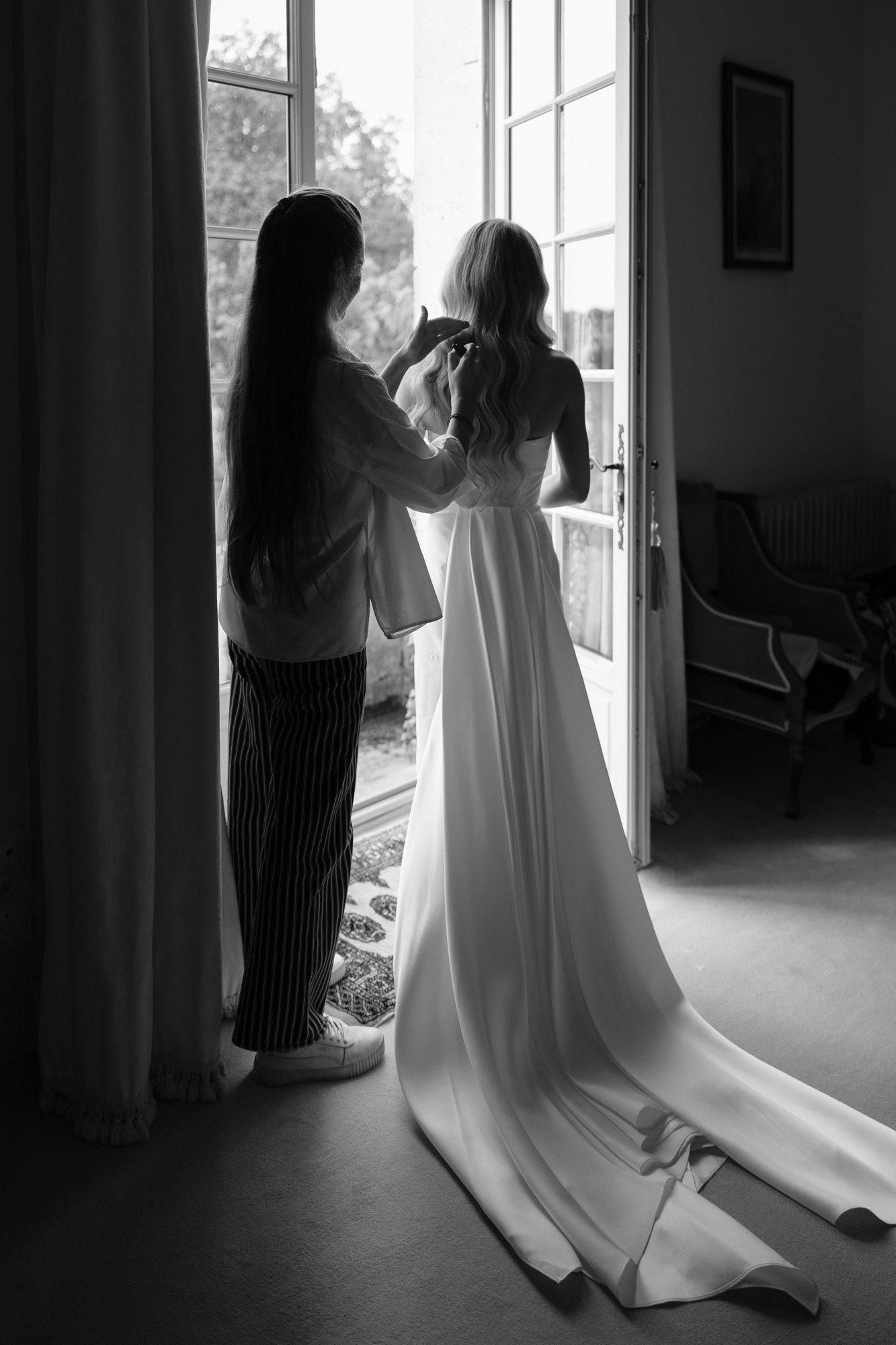 A woman in a long, white gown stands by an open door as another person styles her hair; natural light streams in from outside. Aubeterre sur dronne wedding.