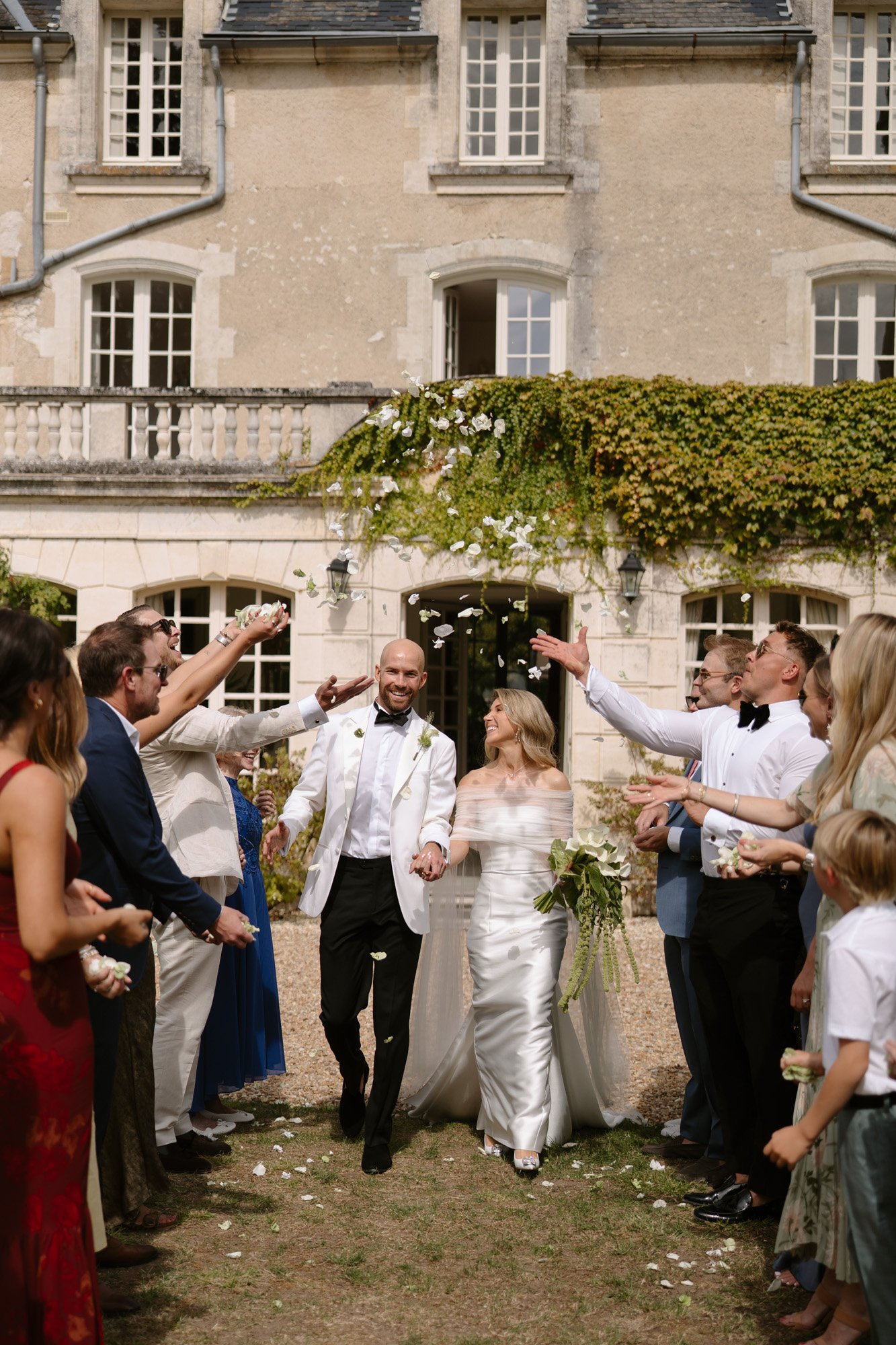 A newlywed couple walks hand in hand outside a stone building as guests throw flower petals and celebrate around them. Aubeterre sur dronne wedding.