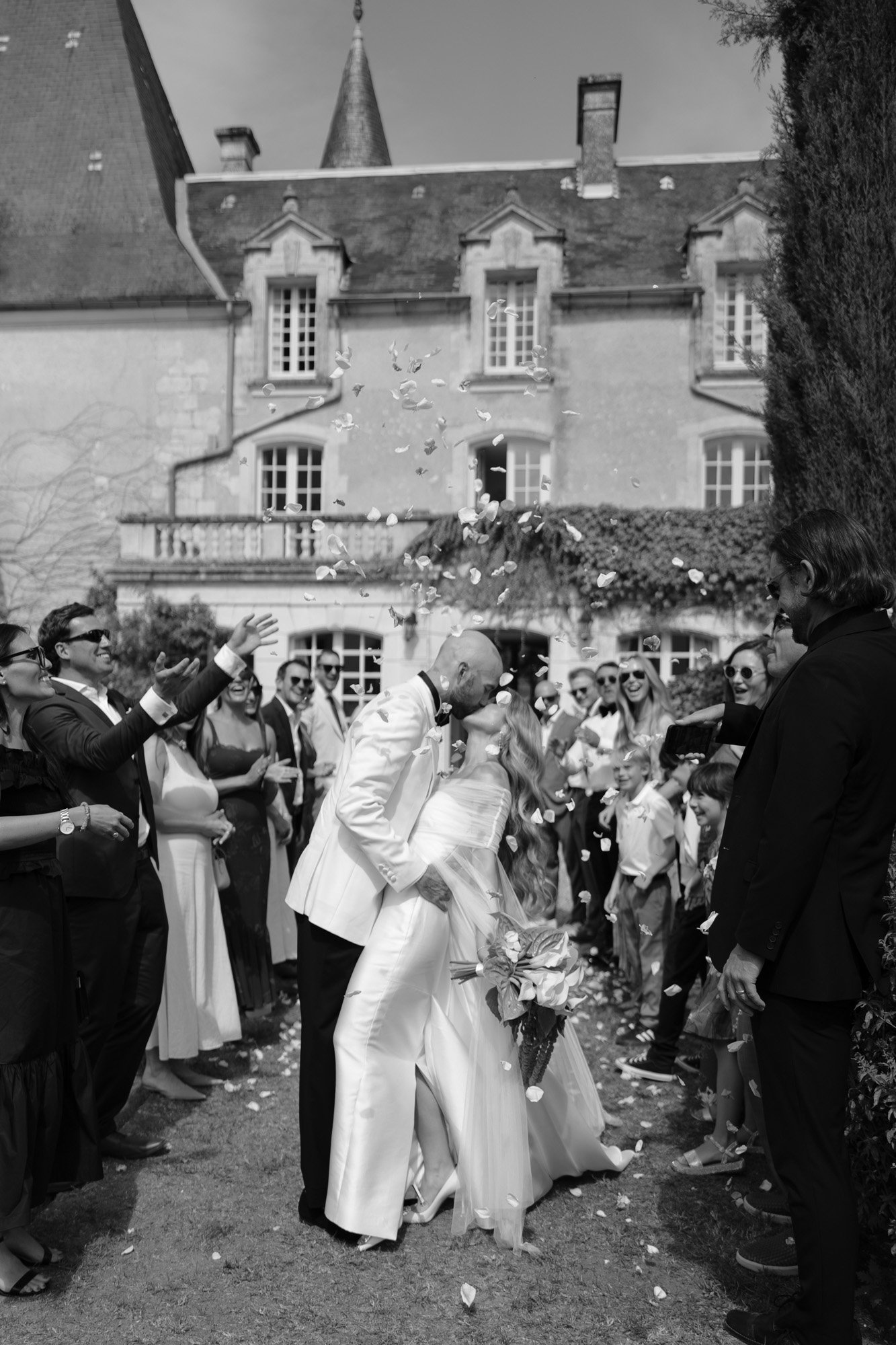 A newlywed couple kisses outdoors while guests throw flower petals; a large stone building stands in the background.
