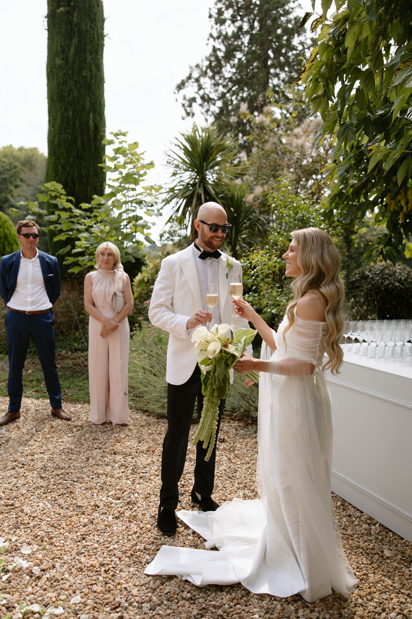 A bride and groom in formal attire toast with champagne outdoors, while two guests stand nearby on a gravel surface, surrounded by greenery. Aubeterre sur dronne wedding.