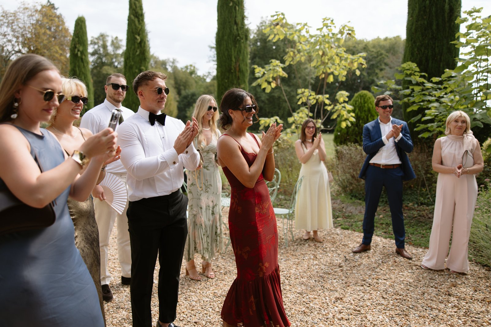 A group of well-dressed people stand outside on a gravel path, clapping and smiling, surrounded by tall trees and greenery. Aubeterre sur dronne wedding.