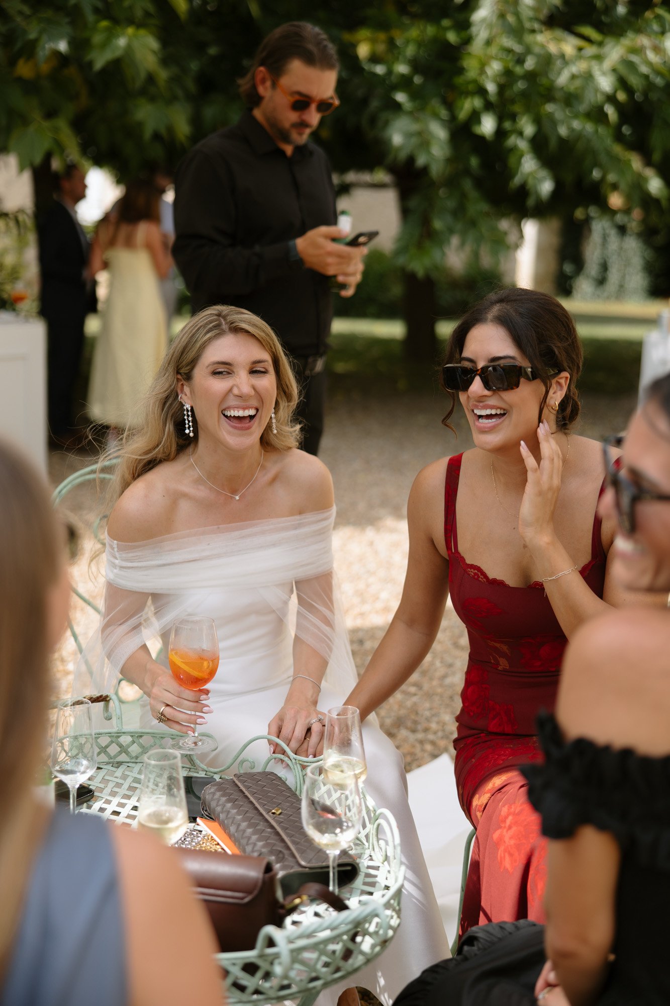 Two women sit at an outdoor table, smiling and laughing. One wears a white dress and holds a drink, the other wears a red dress and sunglasses. Others are visible in the background.