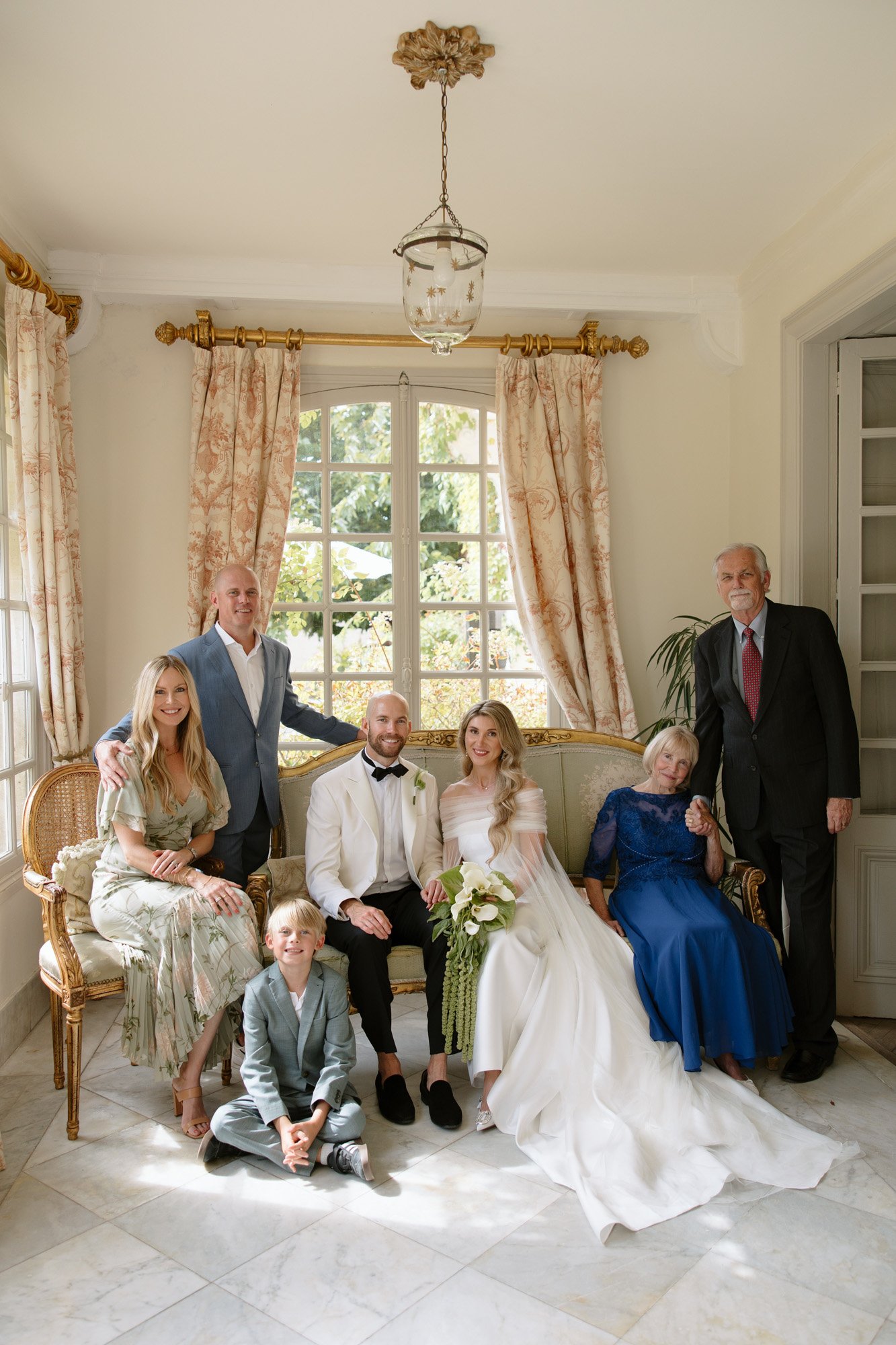 A group of seven people, including a bride and groom, pose together indoors on and around a sofa in a well-lit room with large windows and ornate curtains. Aubeterre sur dronne wedding.