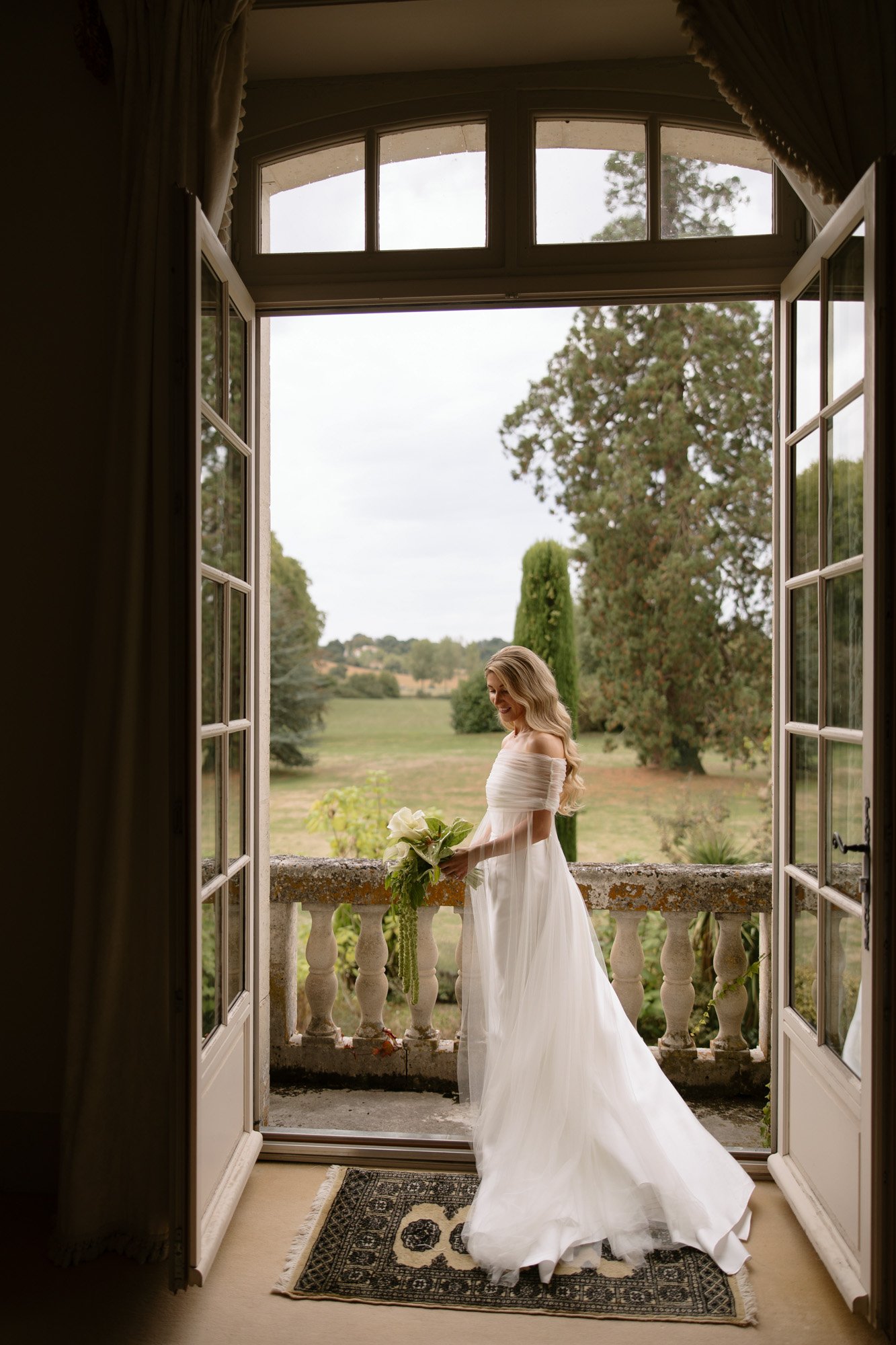 A woman in a white wedding dress stands on a balcony holding a bouquet, looking out over a green landscape through open double doors. Aubeterre sur dronne wedding.