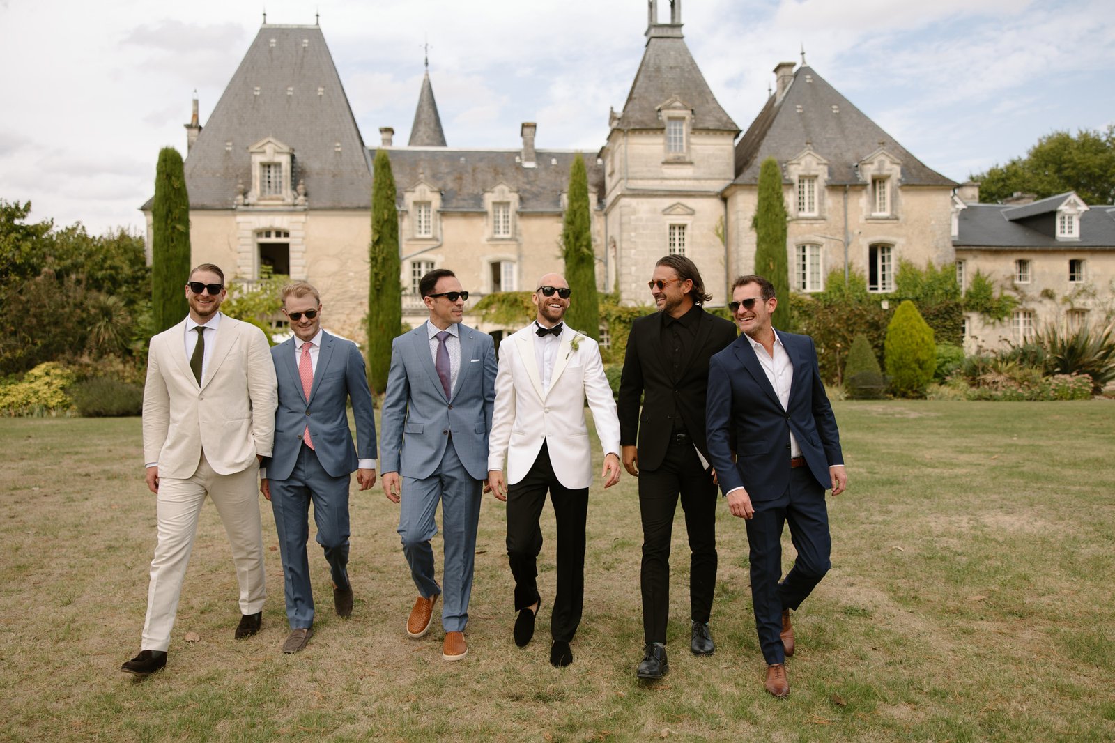 Six men in suits walk on grass in front of a historic chateau with pointed roofs and tall trees, all smiling and wearing sunglasses.