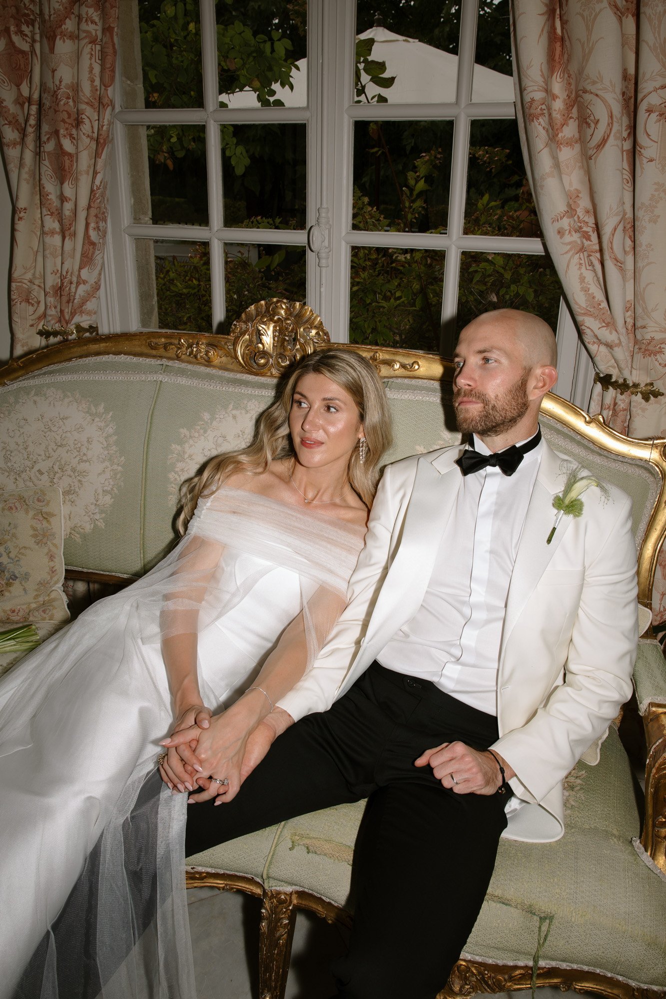A bride and groom in formal attire sit closely together on an ornate sofa, holding hands and looking off camera, with a window and curtains in the background. Aubeterre sur dronne wedding.