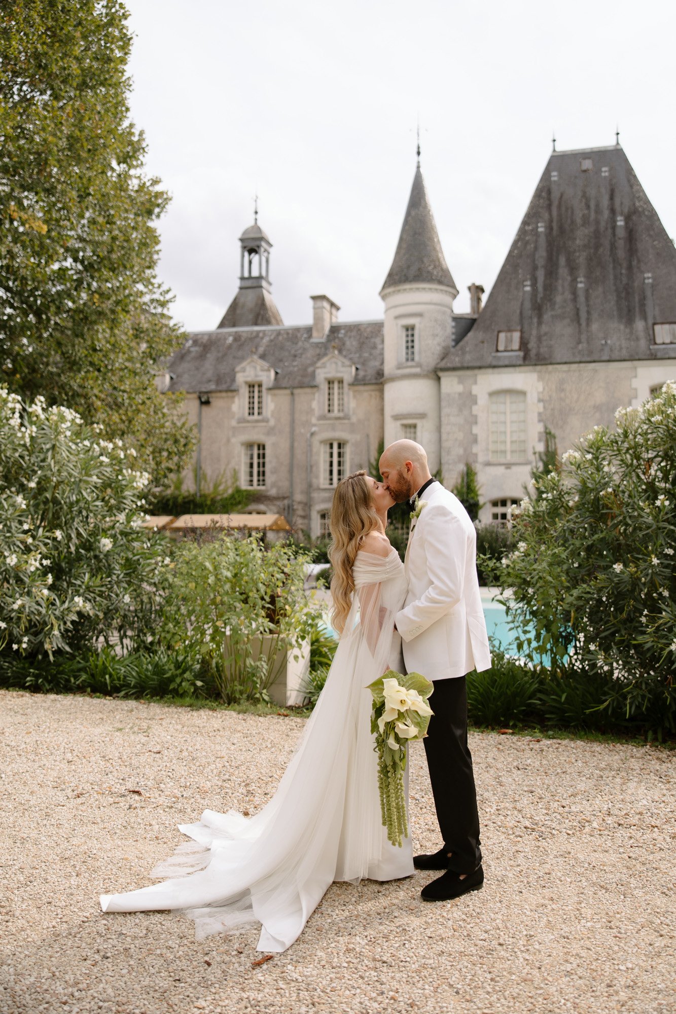 A bride and groom in formal attire share a kiss outdoors in front of a historic stone building, with greenery and a bouquet visible. Aubeterre sur dronne wedding.