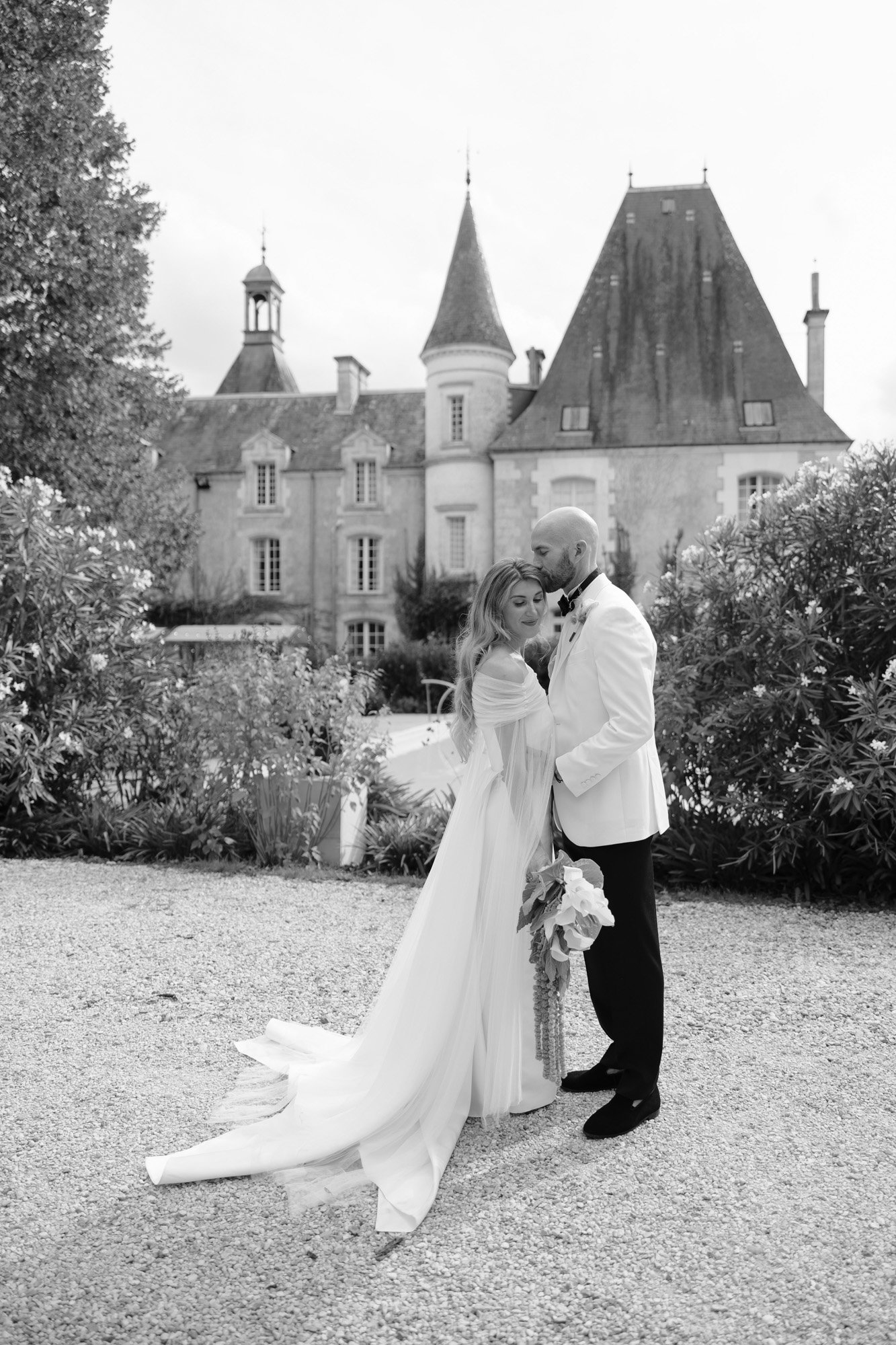 A bride and groom embrace in front of a large historic building with tall peaked roofs, surrounded by greenery. The image is in black and white. Aubeterre sur dronne wedding.