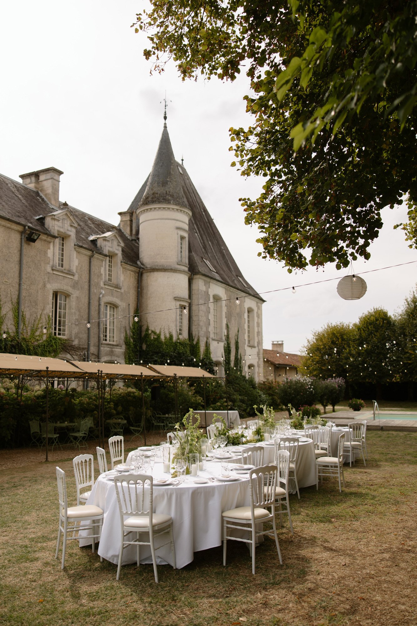 Round tables with white tablecloths and chairs are set for an outdoor event on the lawn of a historic stone building with a conical tower.