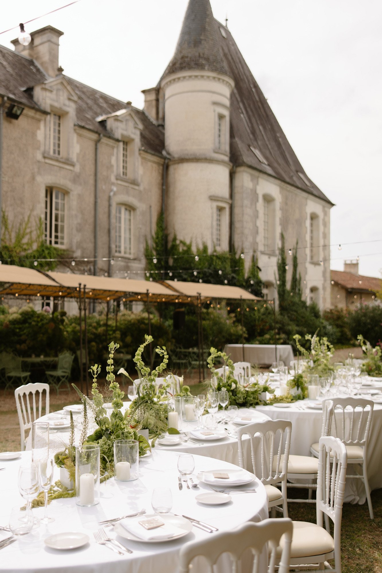 Tables set with white linens and green centerpieces are arranged outdoors in front of an old stone building with a turret. Aubeterre sur dronne wedding.