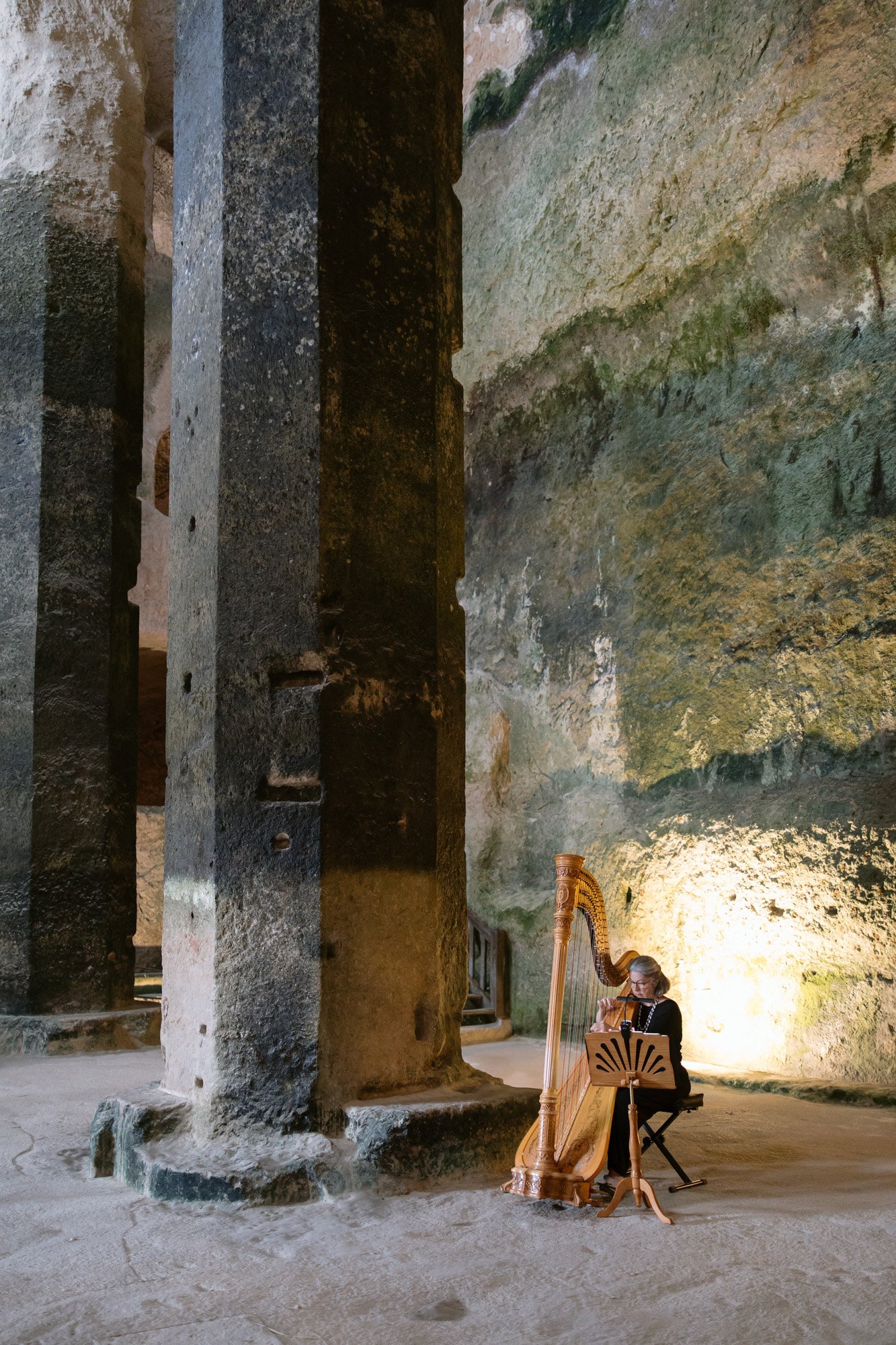 A person plays a harp next to tall stone pillars inside a large, cave-like structure with textured, mossy walls. Aubeterre sur dronne wedding.