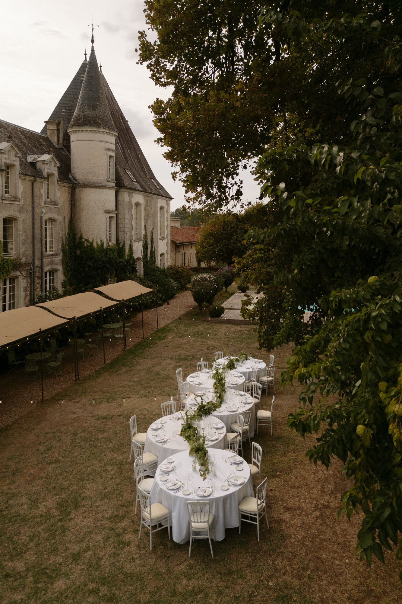Outdoor tables with white tablecloths and chairs are set up on a lawn beside a historic stone building with a tower, under a large tree.