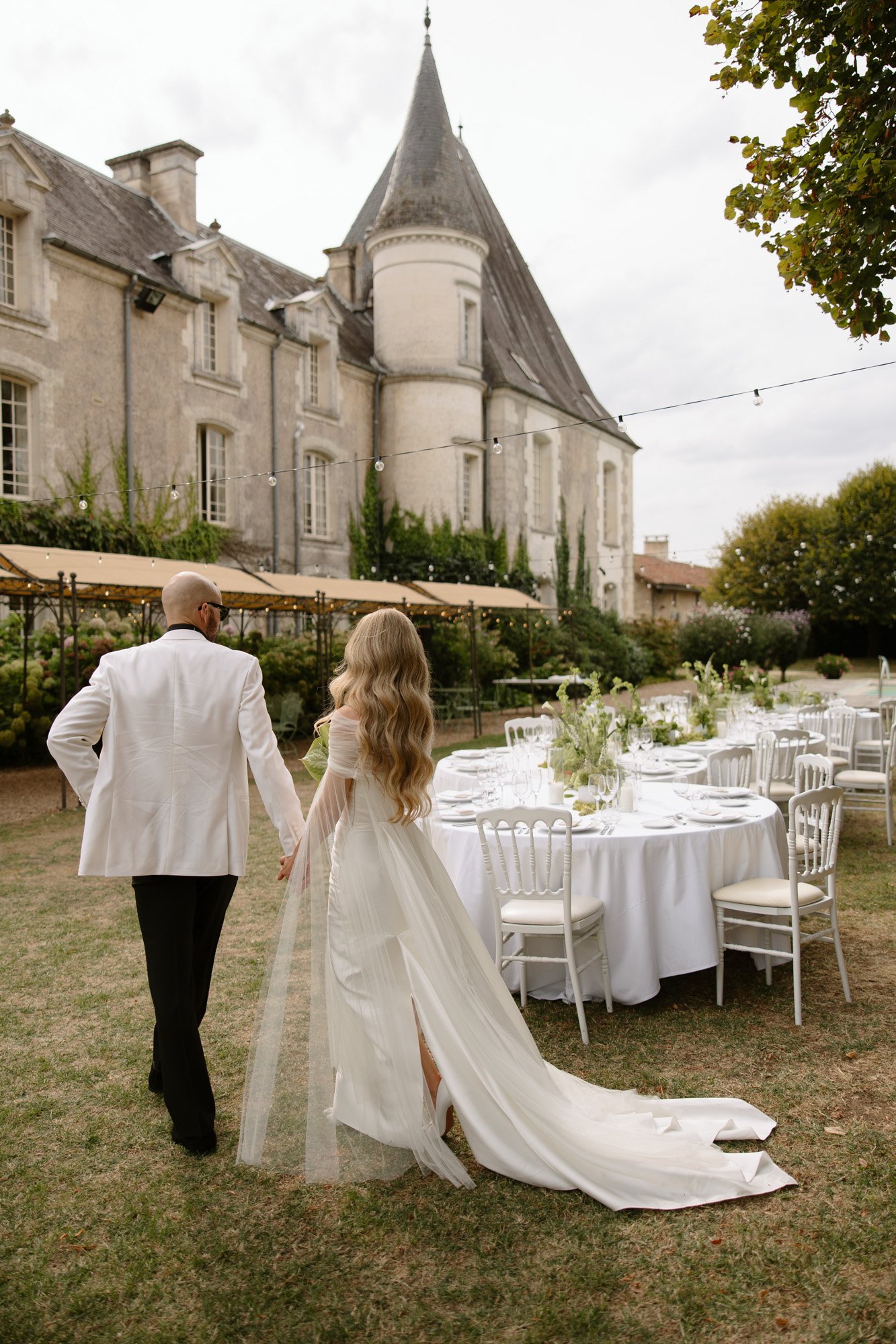 A couple dressed in formal attire walks hand in hand toward round, white-clothed tables set outdoors near a historic stone building. Aubeterre sur dronne wedding.