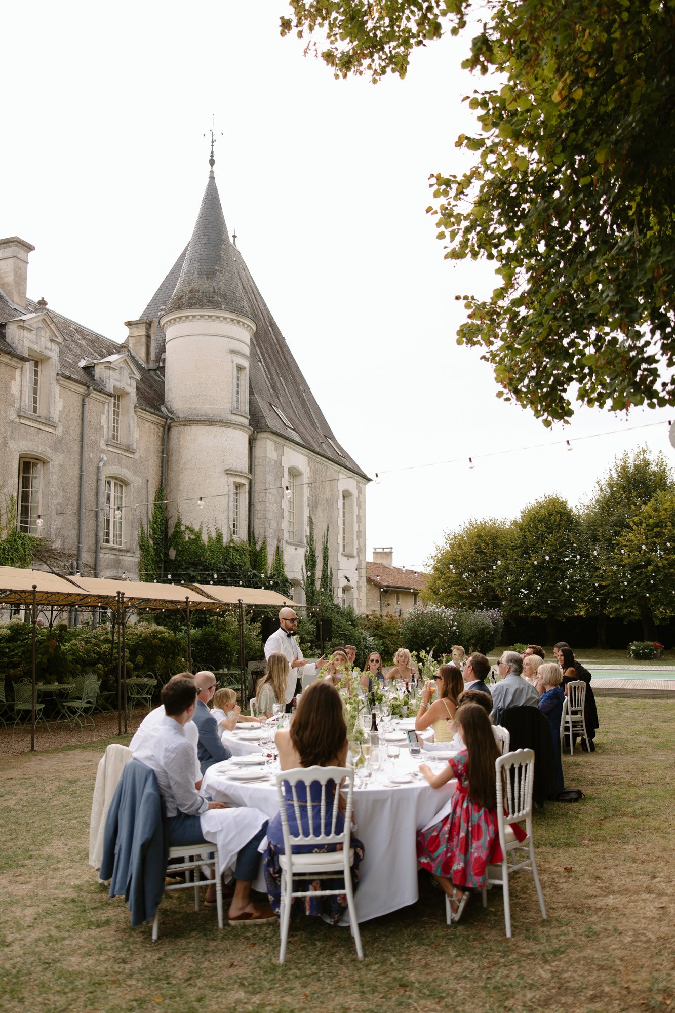 A group of people sit around a long outdoor table set for a meal beside a historic stone building with a tower on a grassy lawn. Aubeterre sur dronne wedding.