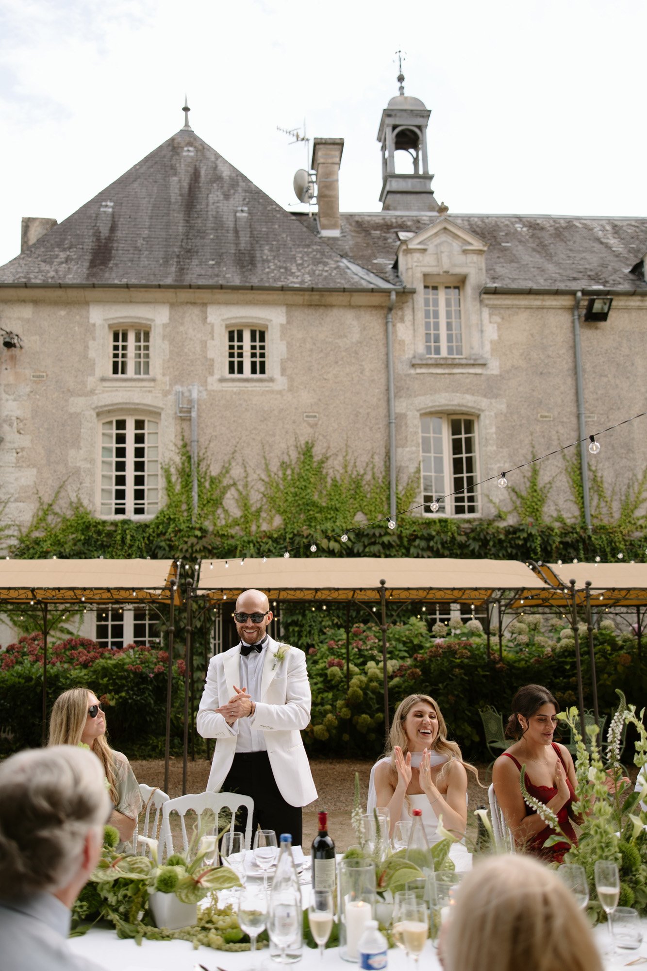 A man in a white jacket stands and addresses seated guests at an outdoor table set for a formal event, with a historic stone building in the background.