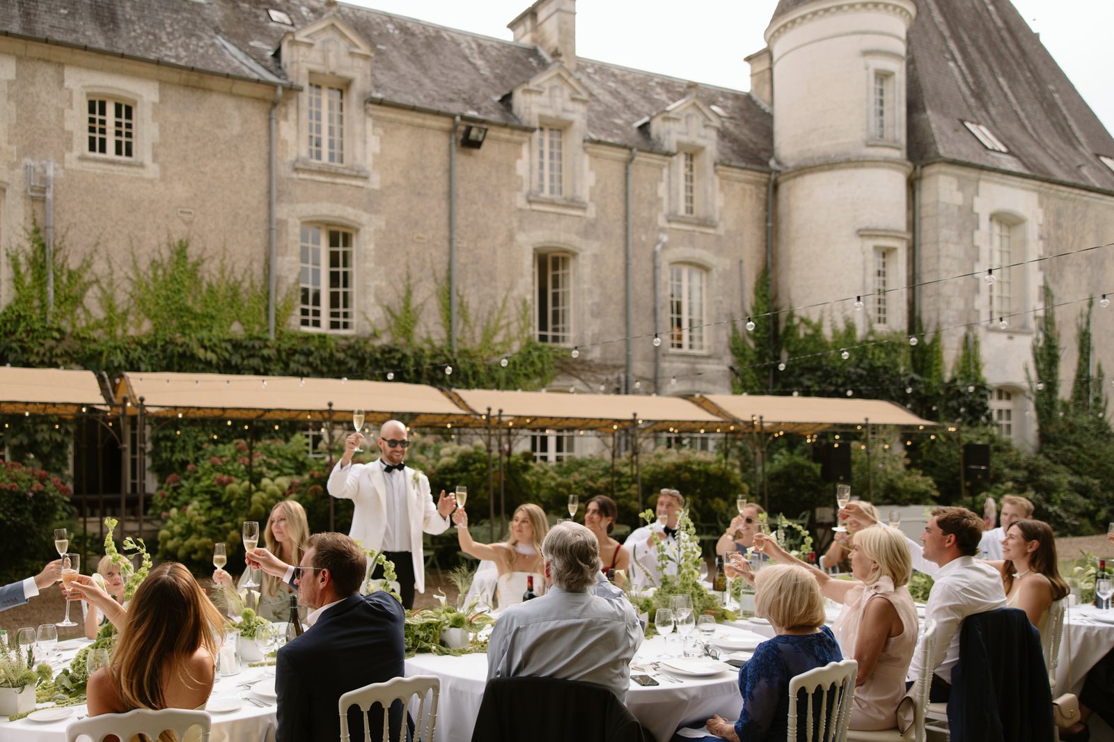 A group of people sit at a long outdoor table set for a formal meal, while a man in a white jacket stands and gives a toast in front of a historic stone building.