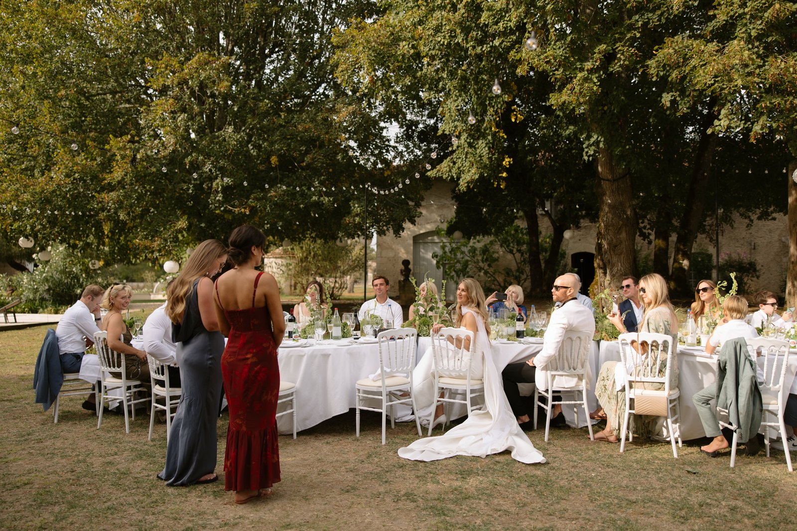 Guests seated at outdoor wedding reception watch as two women stand and speak; the bride in a white gown is seated near the head of the table under trees.