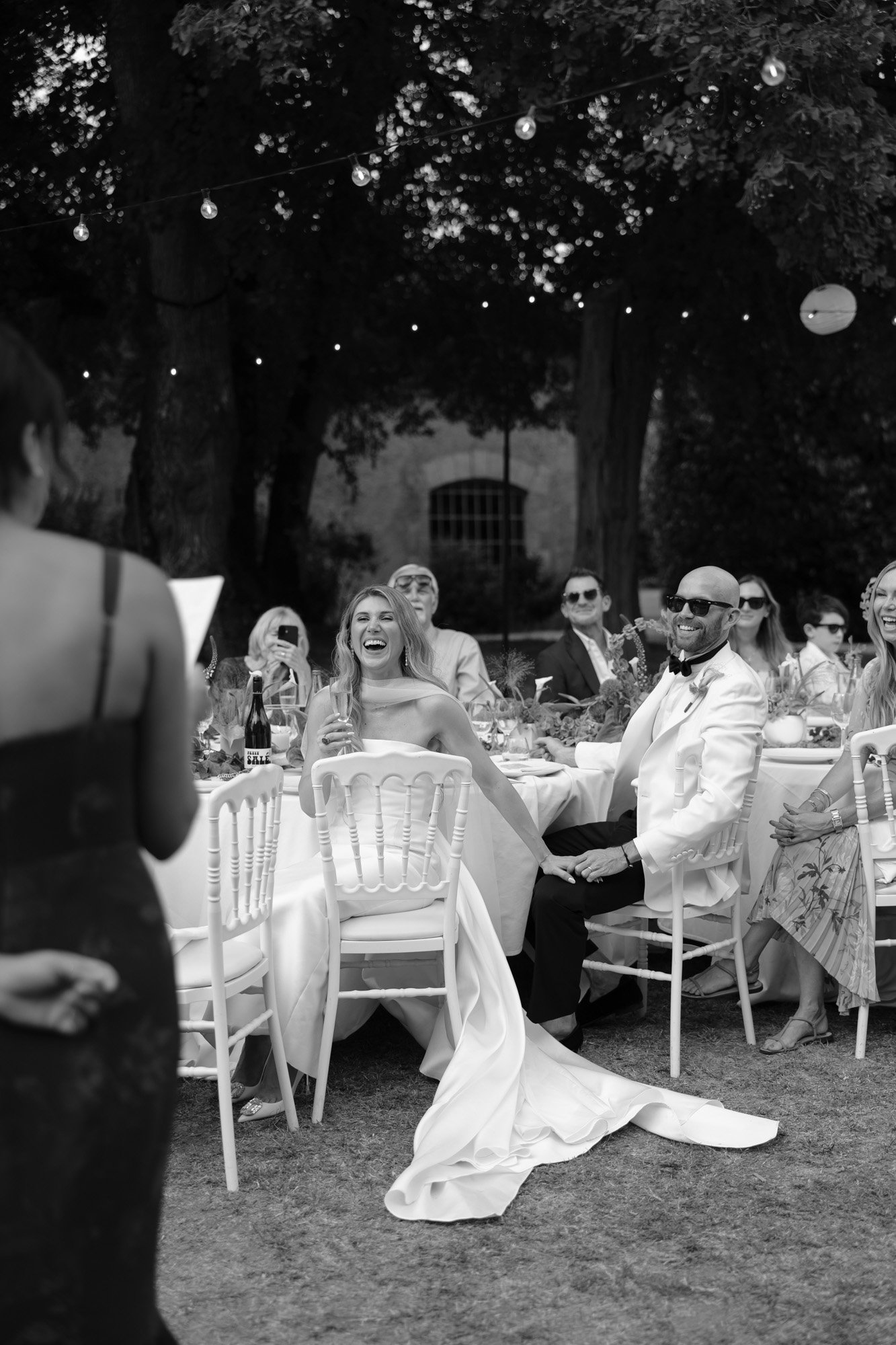 A bride and groom sit at an outdoor reception table, smiling and laughing with guests while listening to a speech.