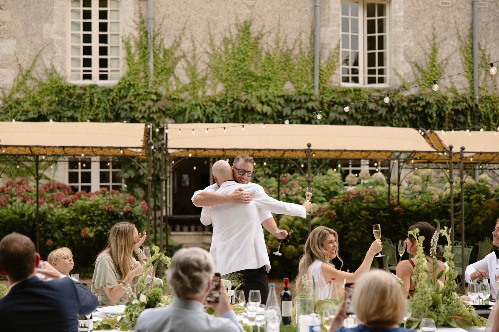 Two people hug at an outdoor event with seated guests watching and celebrating at a long table in front of a vine-covered building.