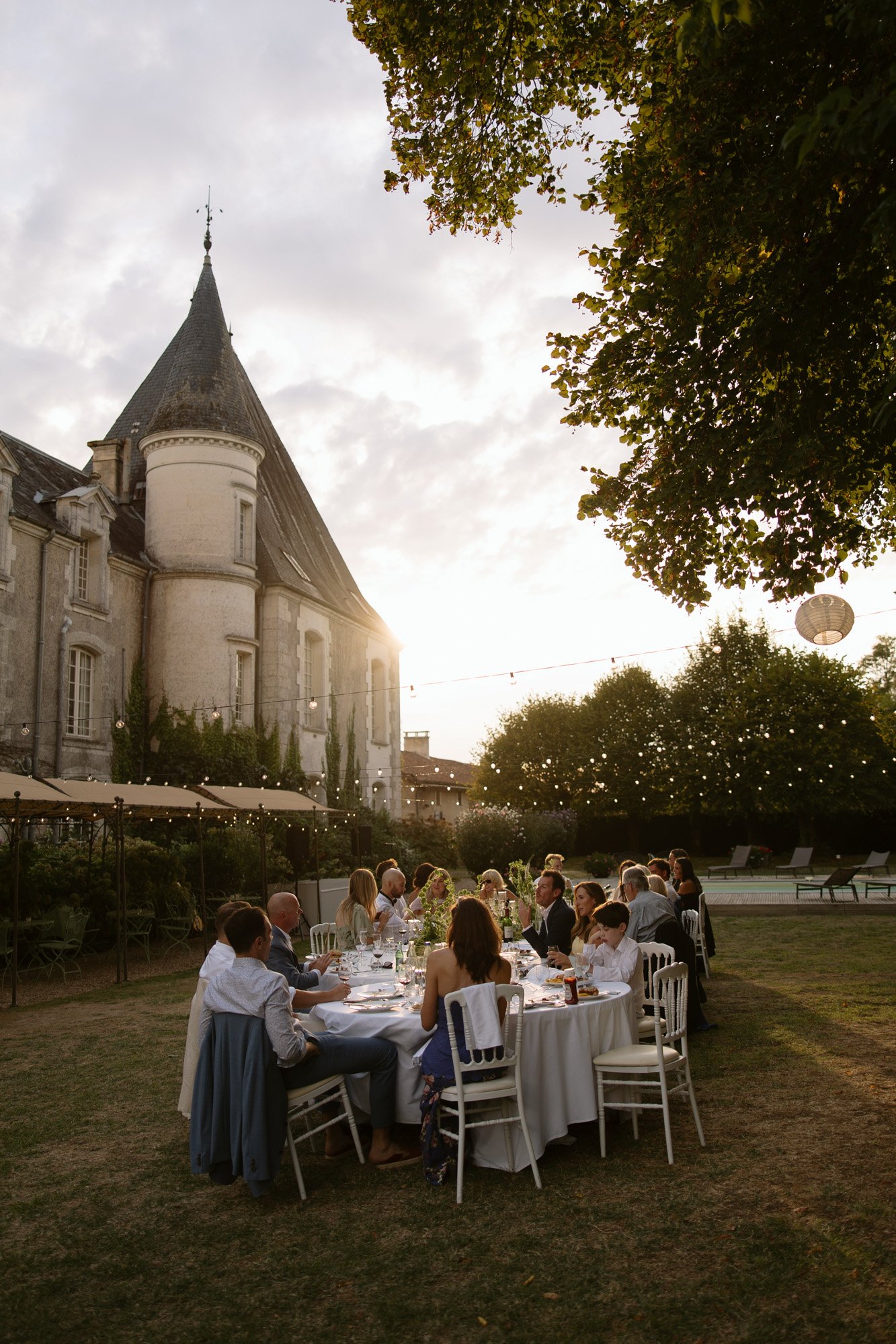 A group of people sit around a dinner table outdoors near a historic stone building at sunset, with string lights and trees in the background.