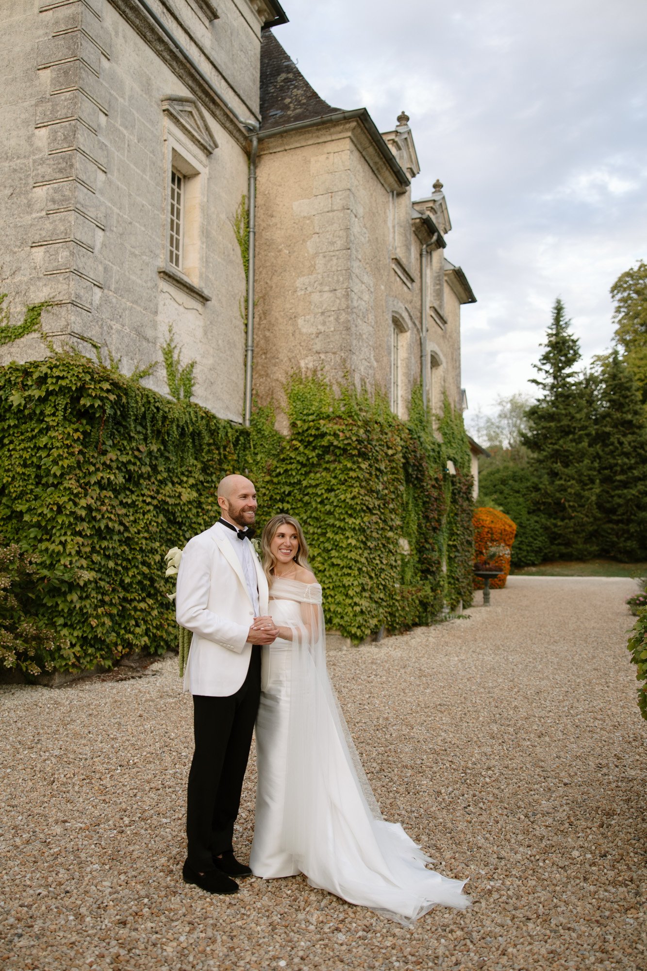 A bride and groom stand together outside a large stone building covered in ivy, posing for a wedding photo on a gravel pathway.