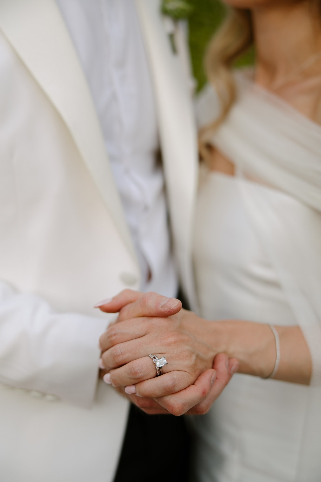 Two people in white formal attire hold hands, with one person wearing a ring with a large stone on their finger.